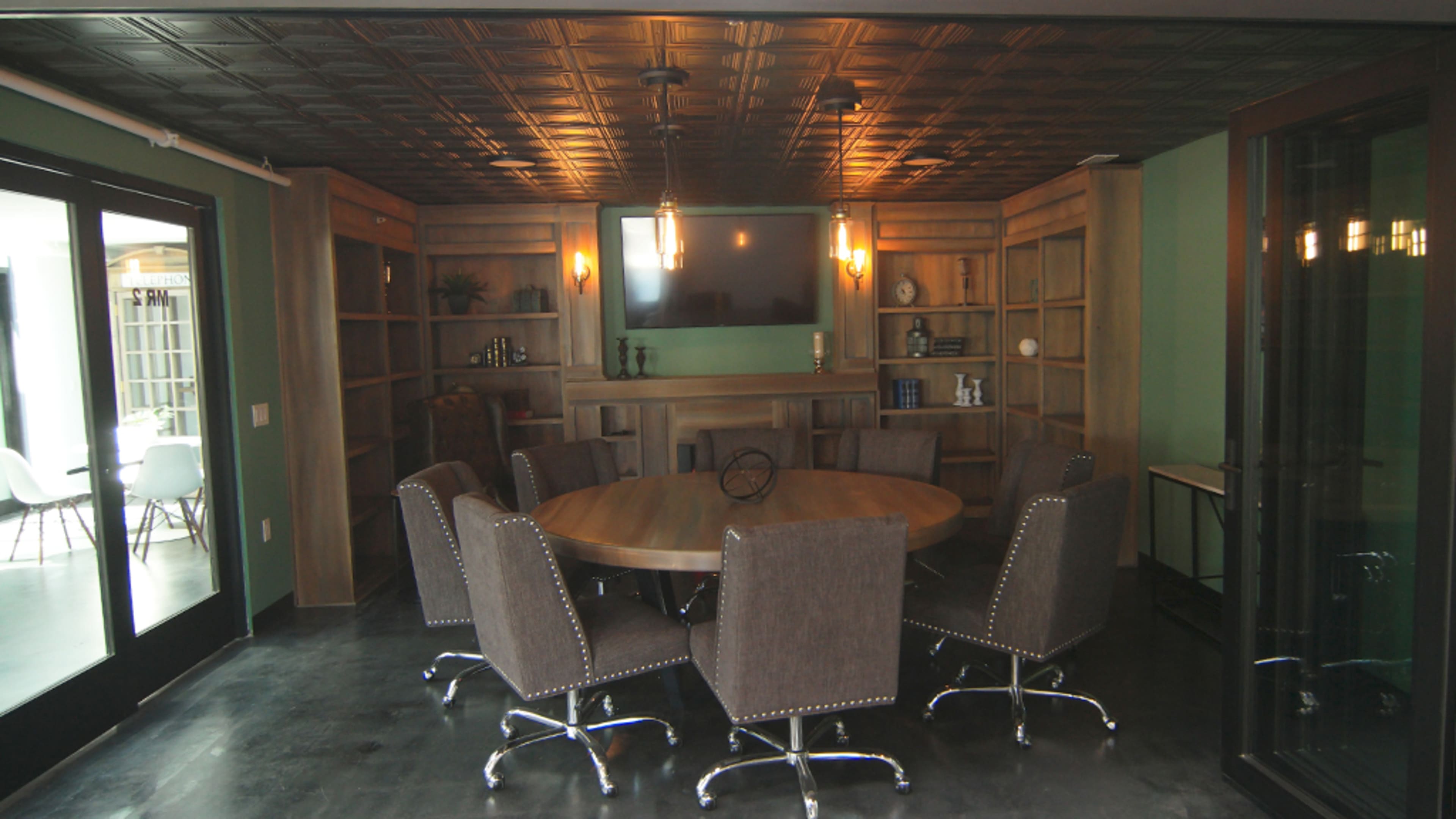 A circular table surrounded by six upholstered chairs is set in a well-lit meeting room featuring wood-paneled shelves and a large screen.