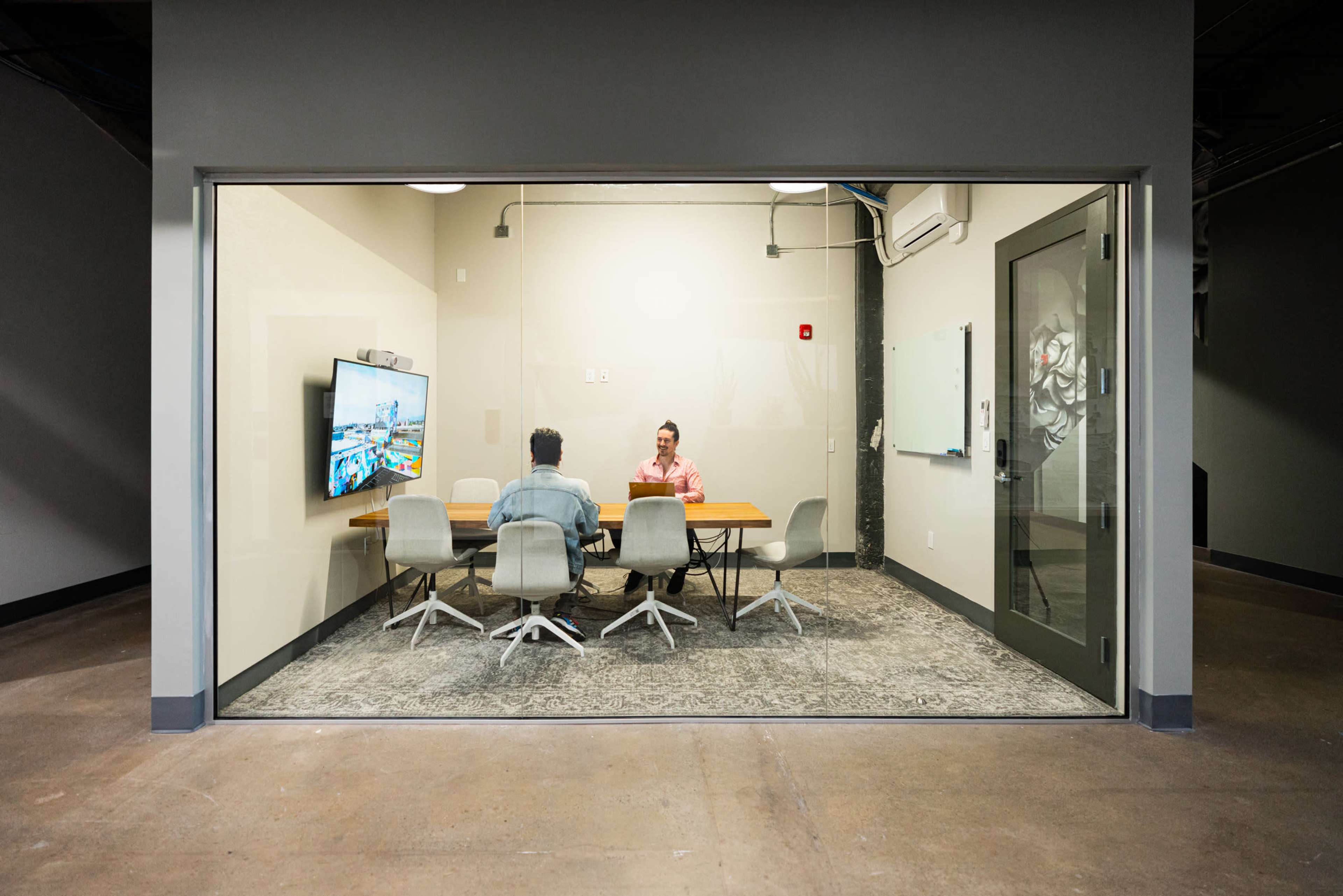 Two people are seated at a conference table in a glass-walled meeting room, with a large screen displaying a digital presentation.