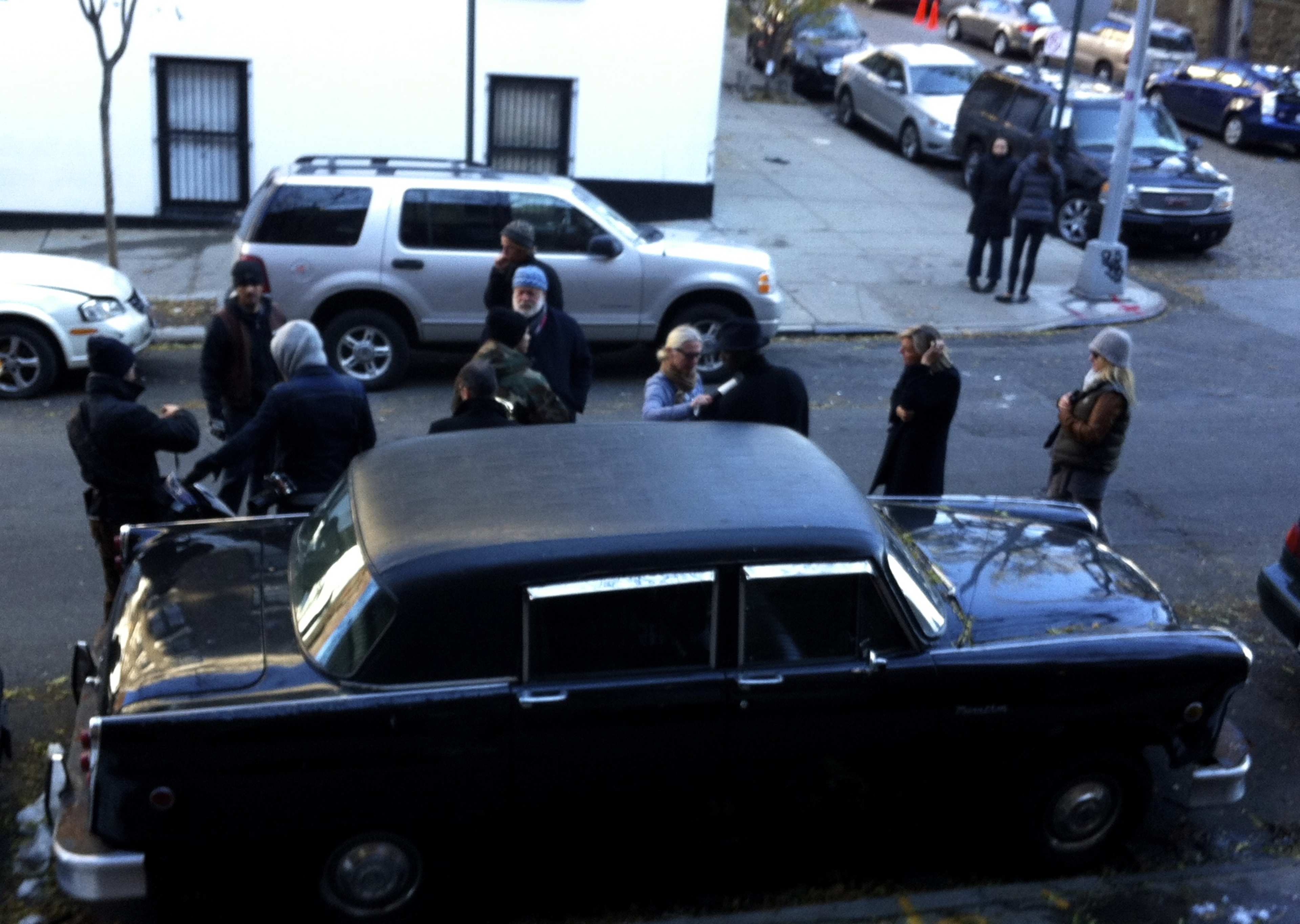 A group of people gathers around a black vintage car on a city street with parked vehicles in the background.