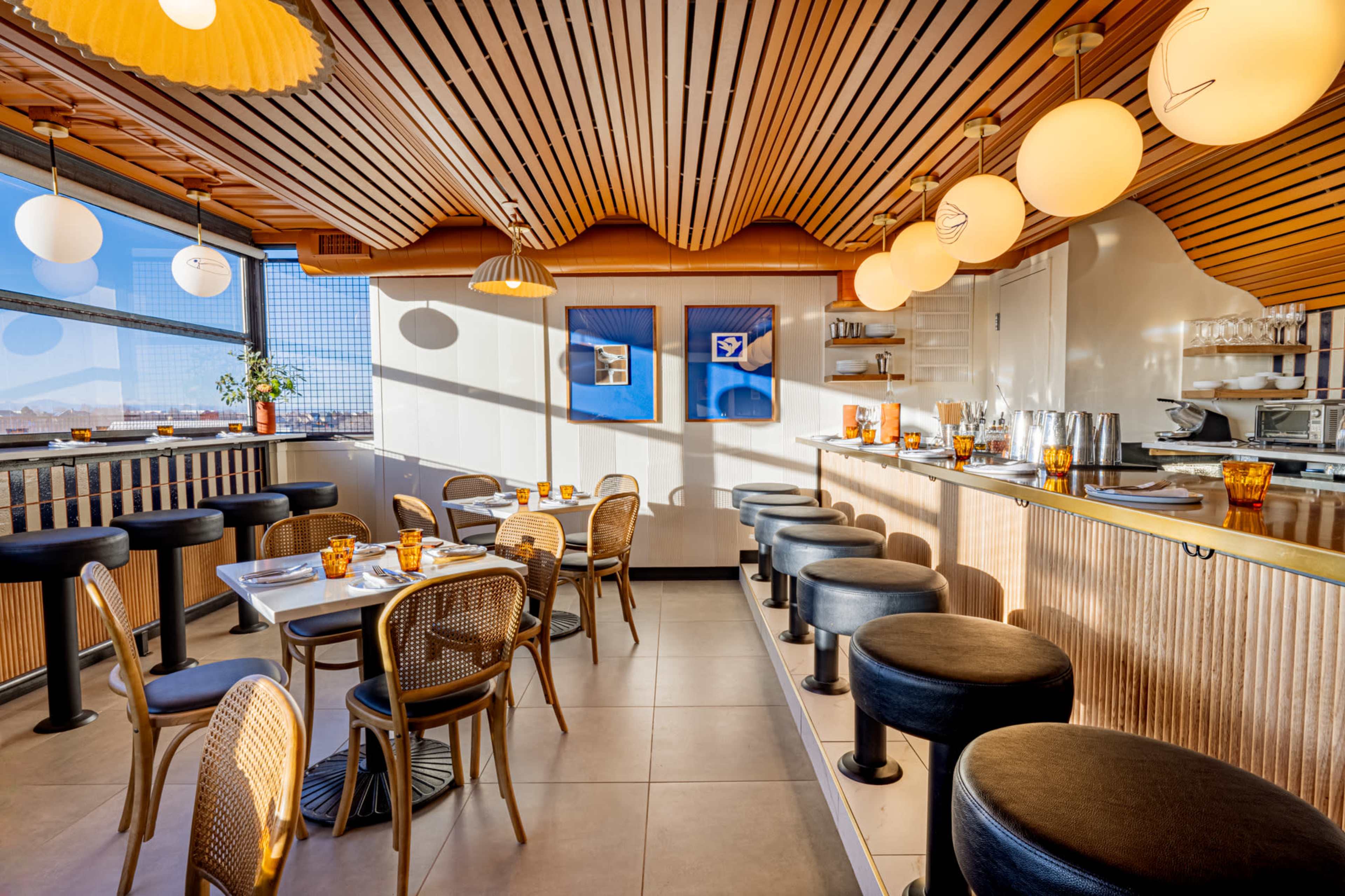 The image shows a modern restaurant interior with wooden accents, round light fixtures, and a mix of round and square tables set for dining.