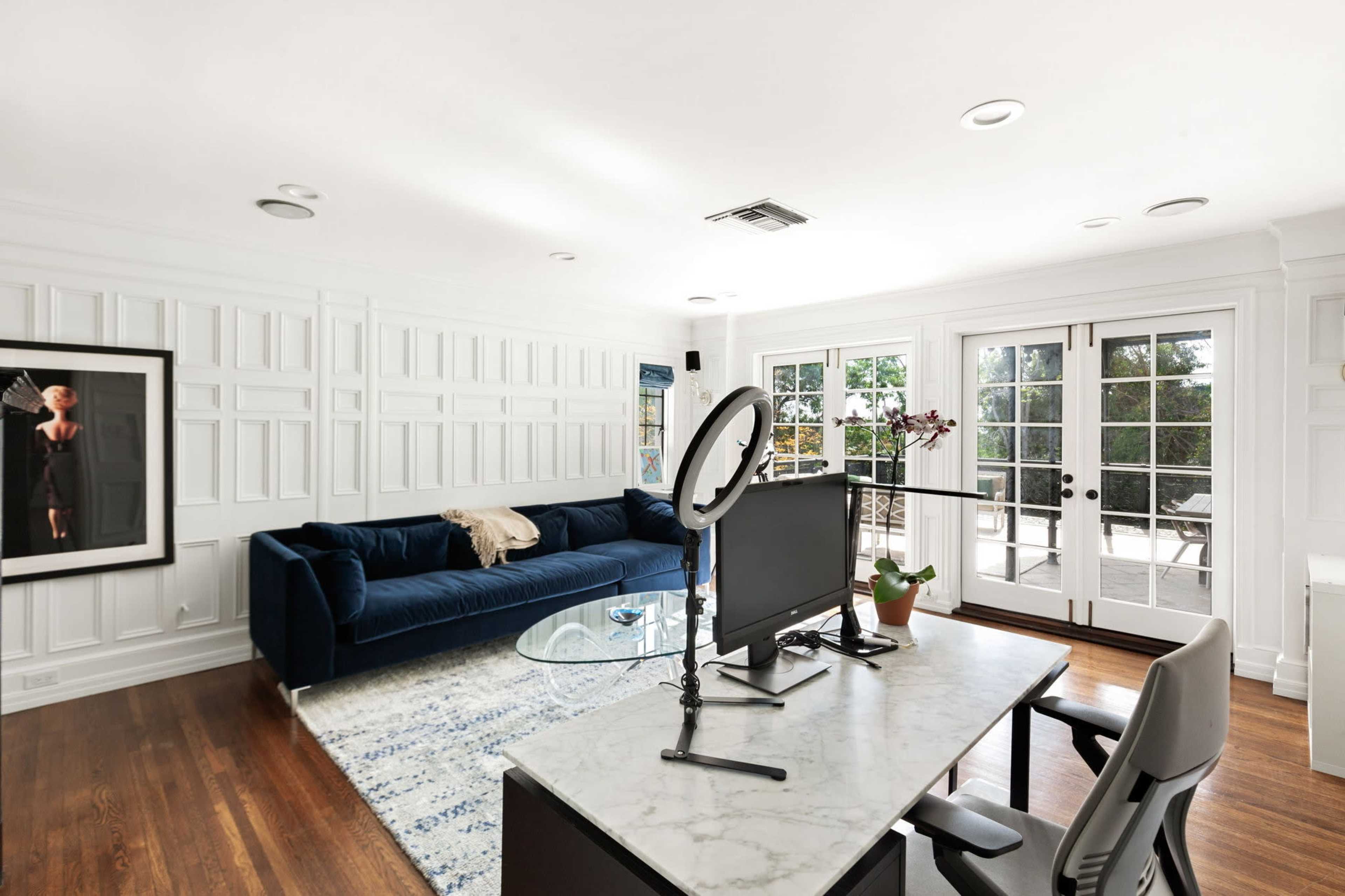 The image shows a well-lit living room featuring a blue velvet sofa, a glass coffee table, and a workspace with a monitor and chair, along with large glass doors leading to a patio.