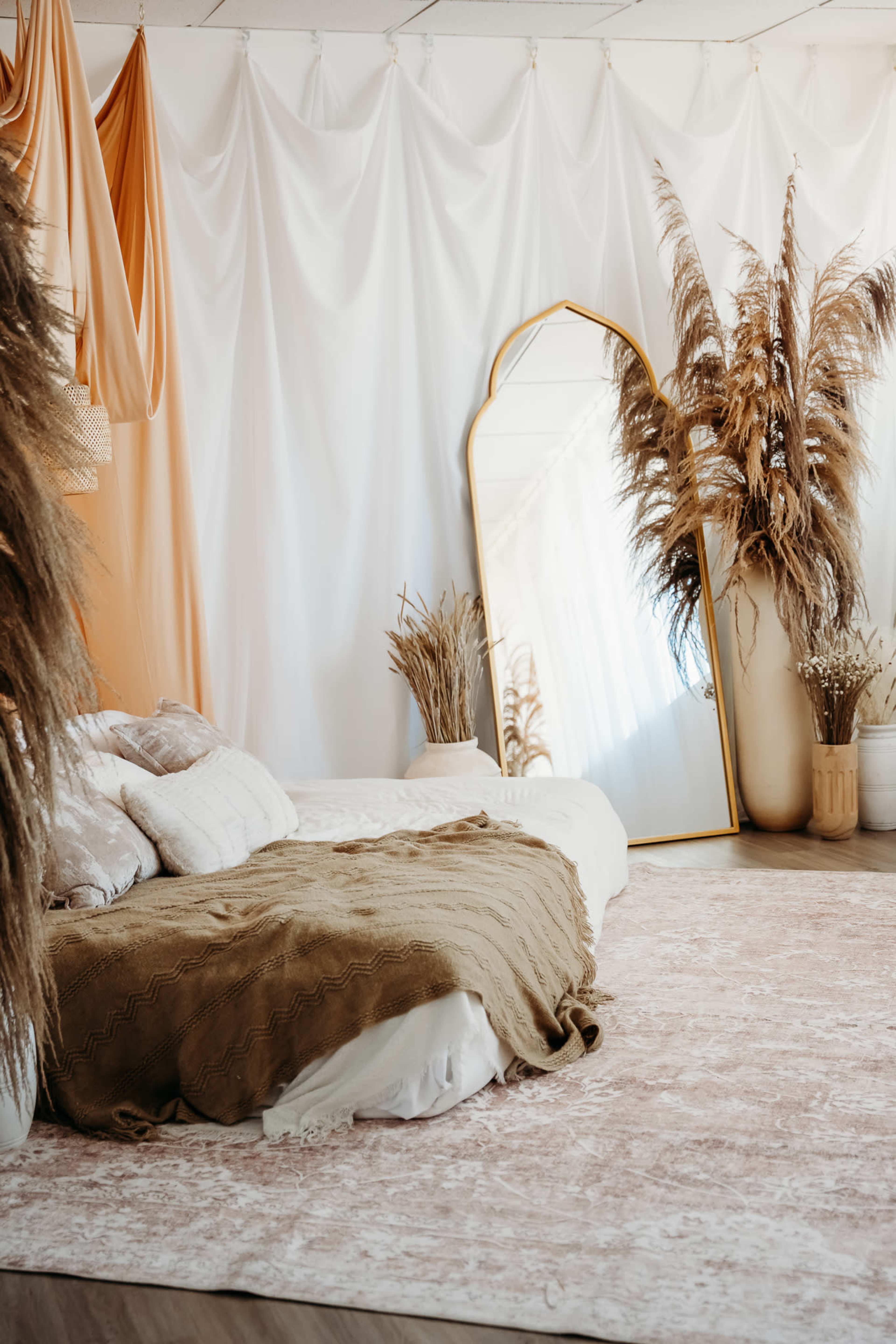 A cozy bedroom features a bed with a textured blanket, a large mirror, and decorative plants arranged beside a softly colored rug.