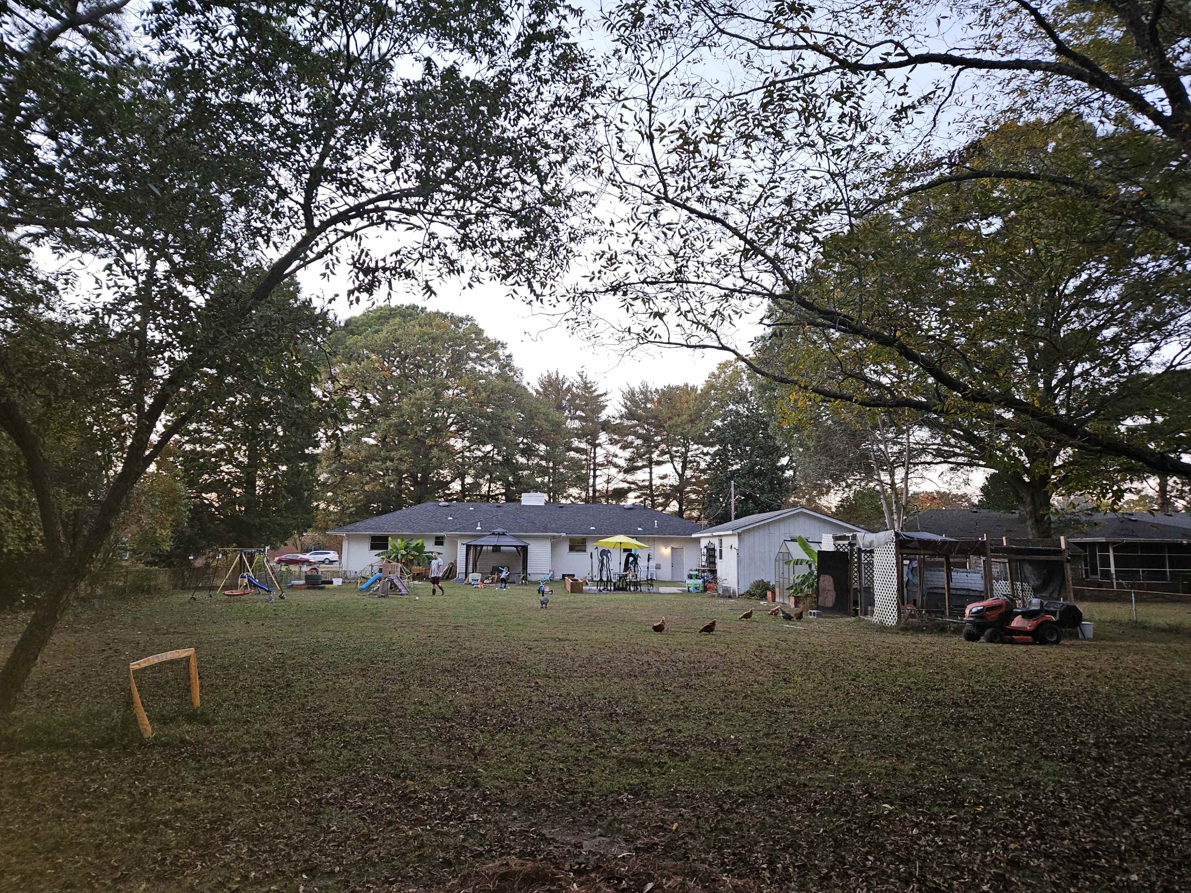 A backyard scene features a house surrounded by trees, with a playground set, chickens, and a lawnmower present in the yard.