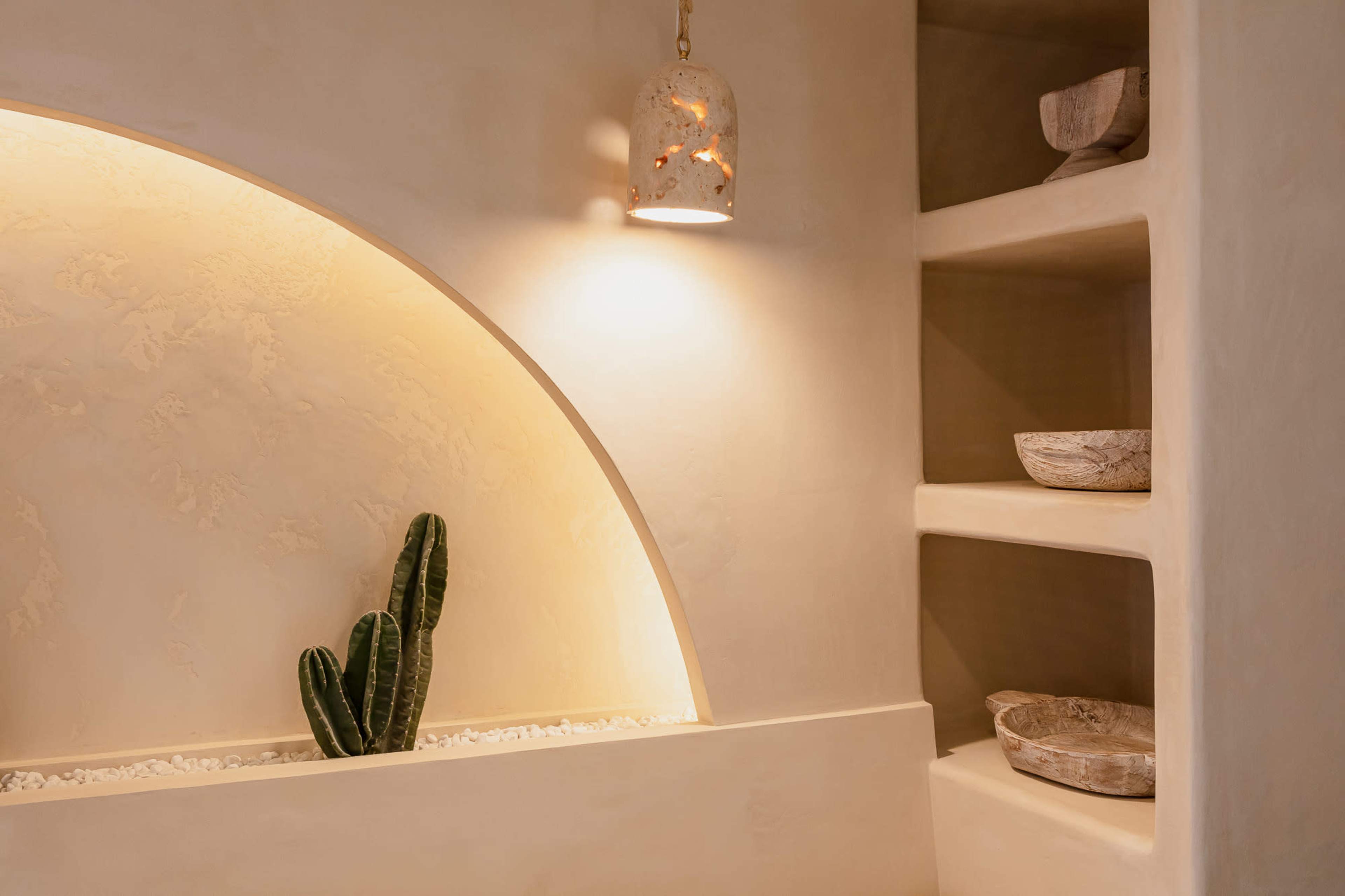 A clean, minimalist interior features a curved alcove with a lit wall niche containing a plant, complemented by shelves of wooden bowls.