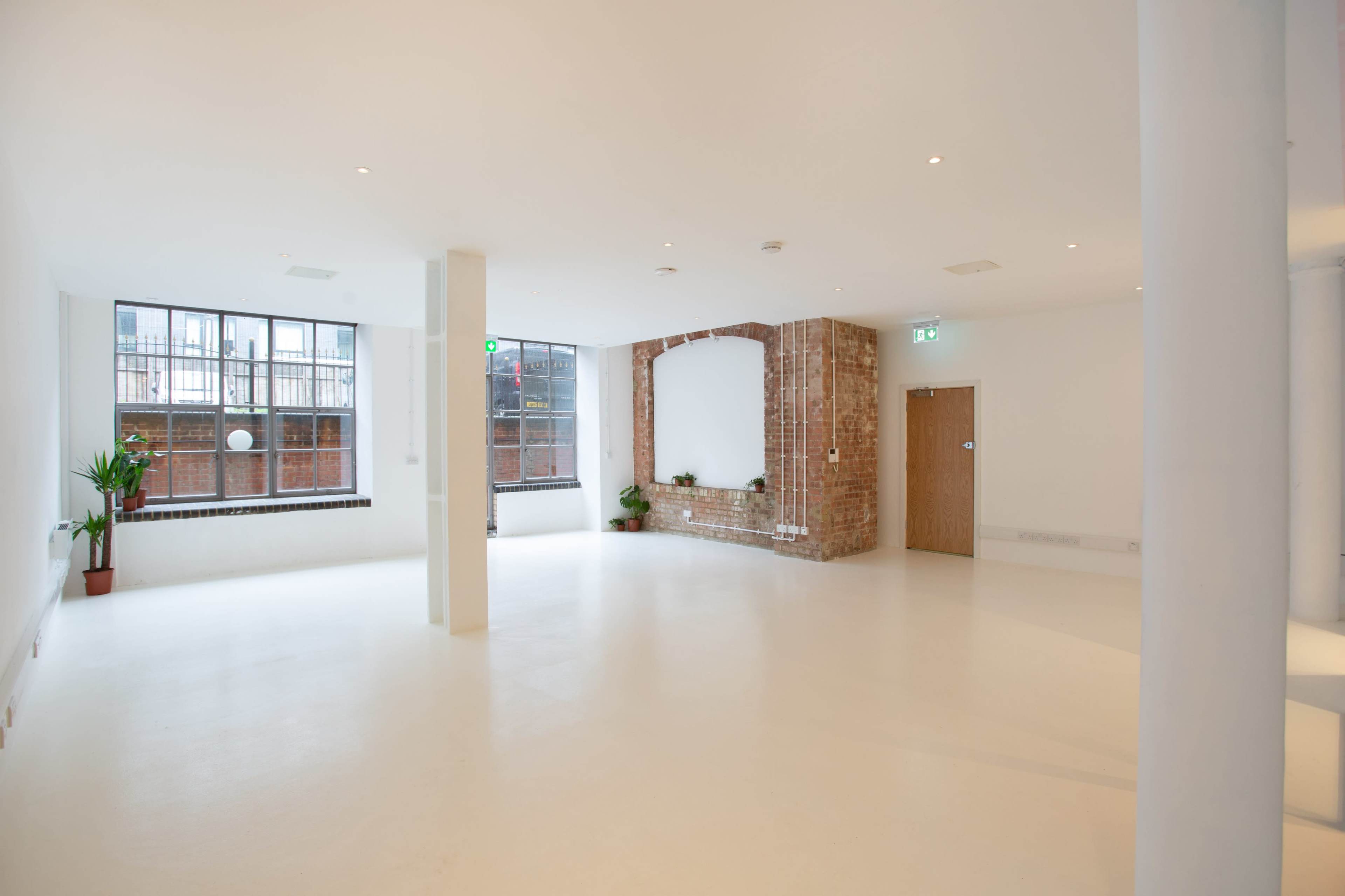 A spacious, empty room with white walls, a brick accent wall, large windows, and minimal furniture.