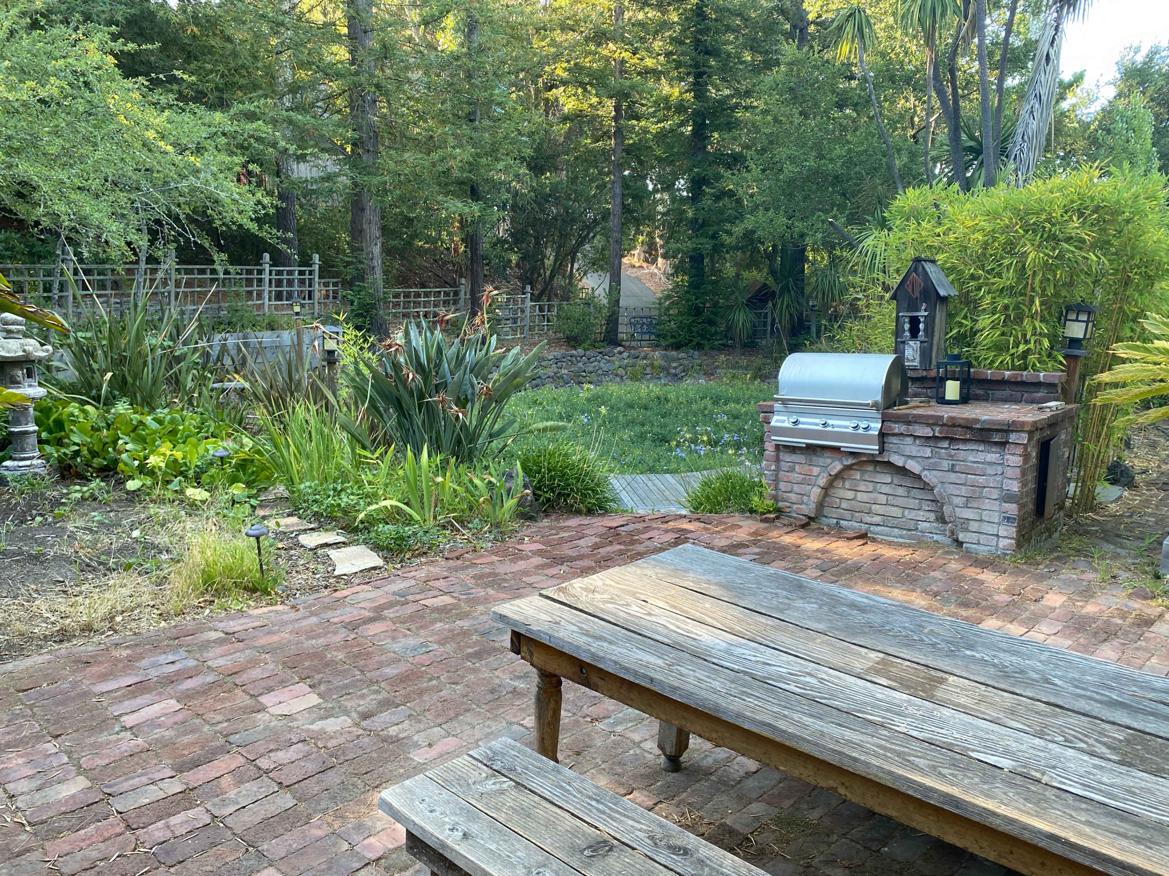 A backyard scene featuring a brick patio with a wooden table and chairs, a stone barbecue grill, and lush greenery in the background.