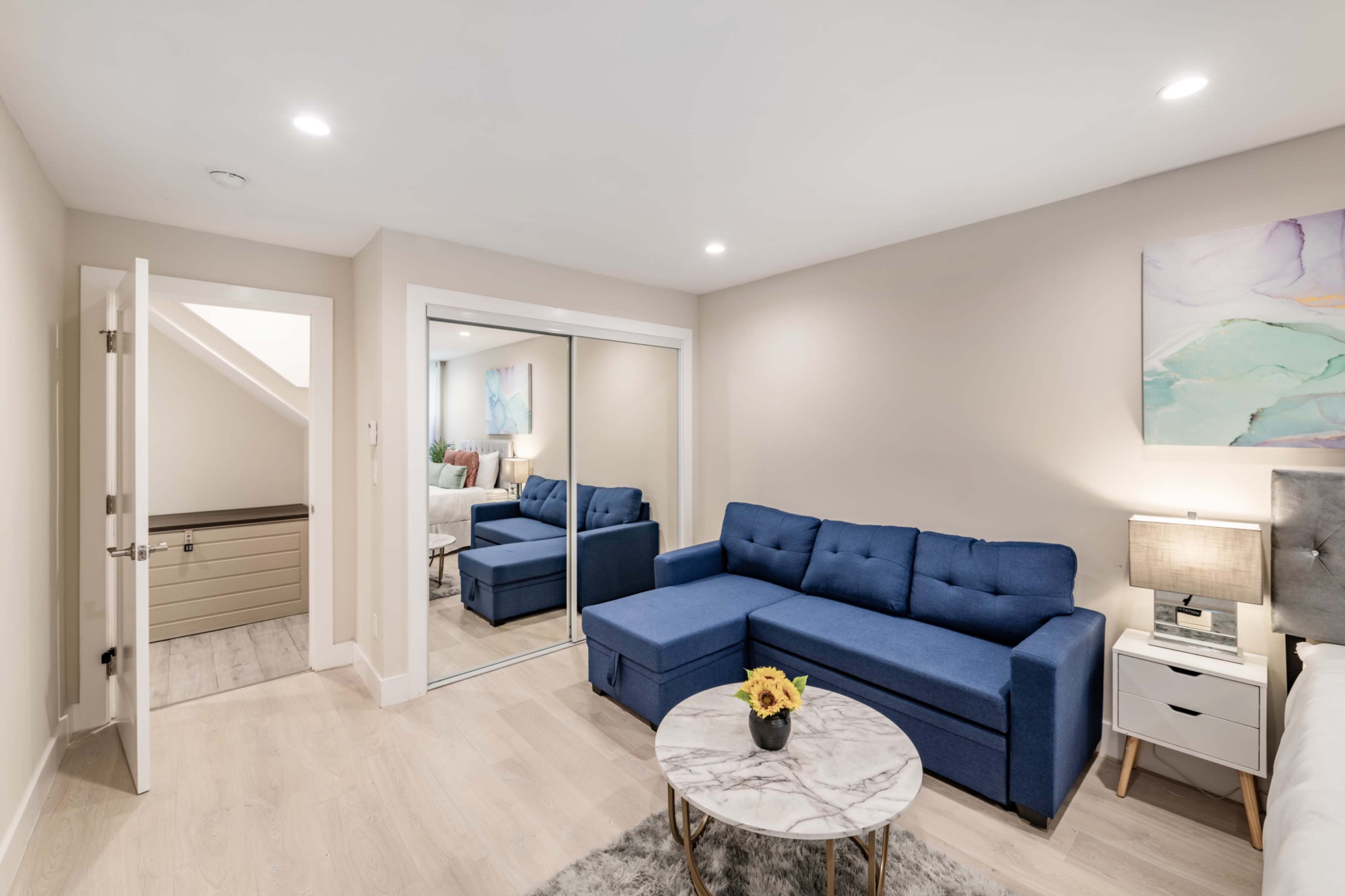 The image shows a modern living space featuring a blue sectional sofa, a round marble coffee table, a mirrored closet, and soft lighting.