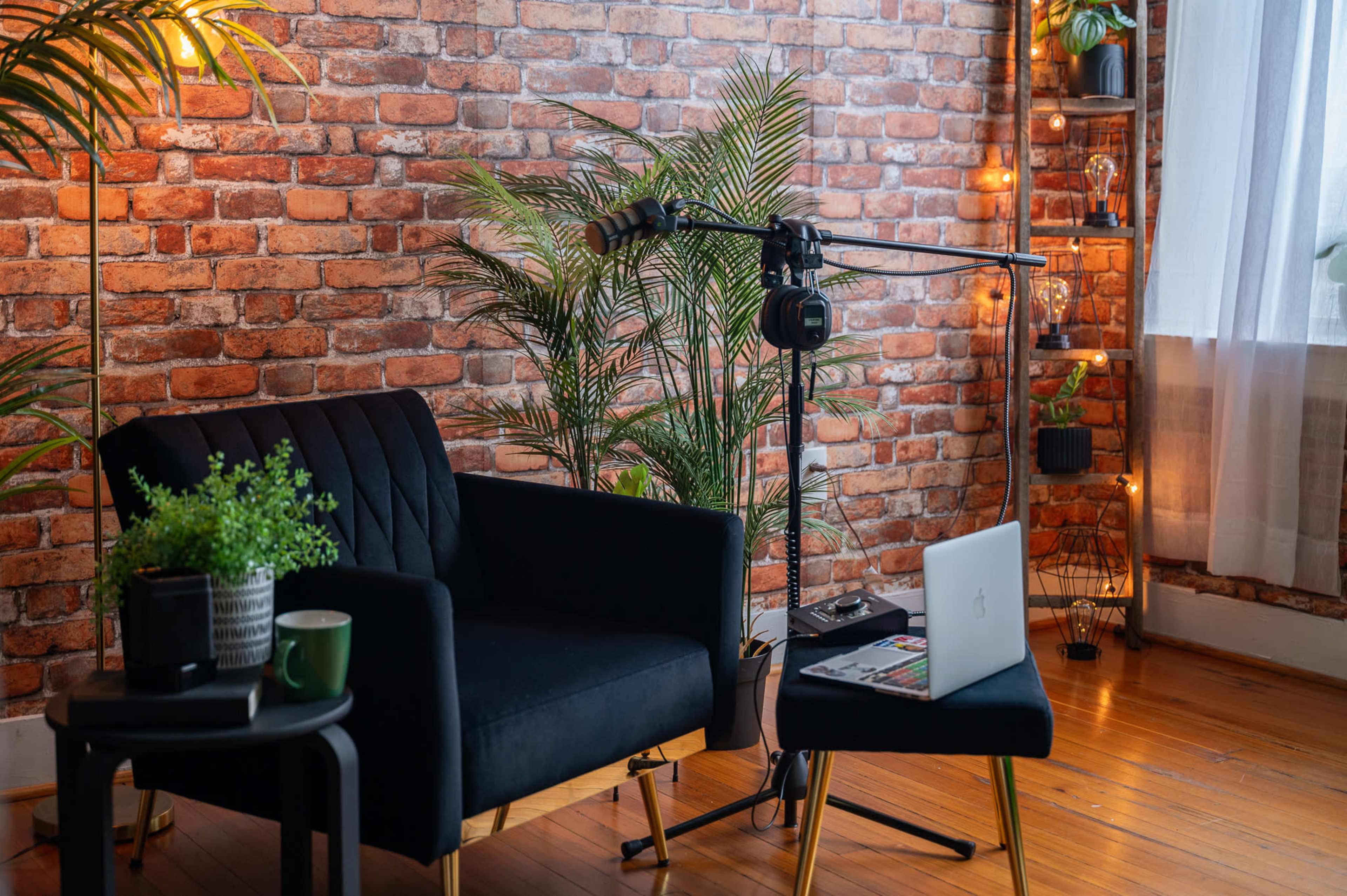 A stylish room with a black armchair, a small table with a green plant and a cup, and a laptop beside a microphone setup against a brick wall.