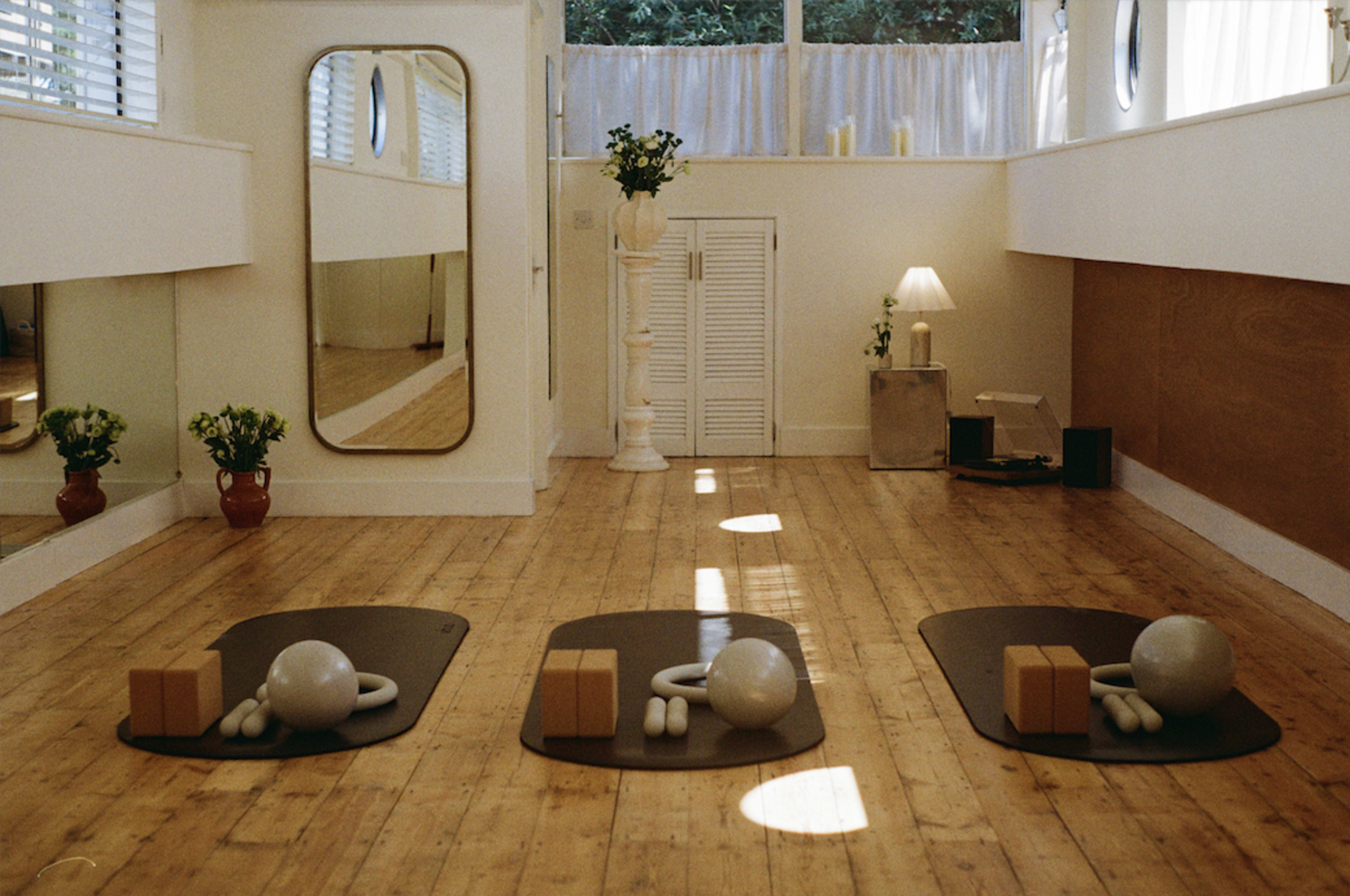 The image shows a bright, minimalist yoga studio with three exercise mats, various props like weights and wooden blocks, and natural light streaming through large windows.