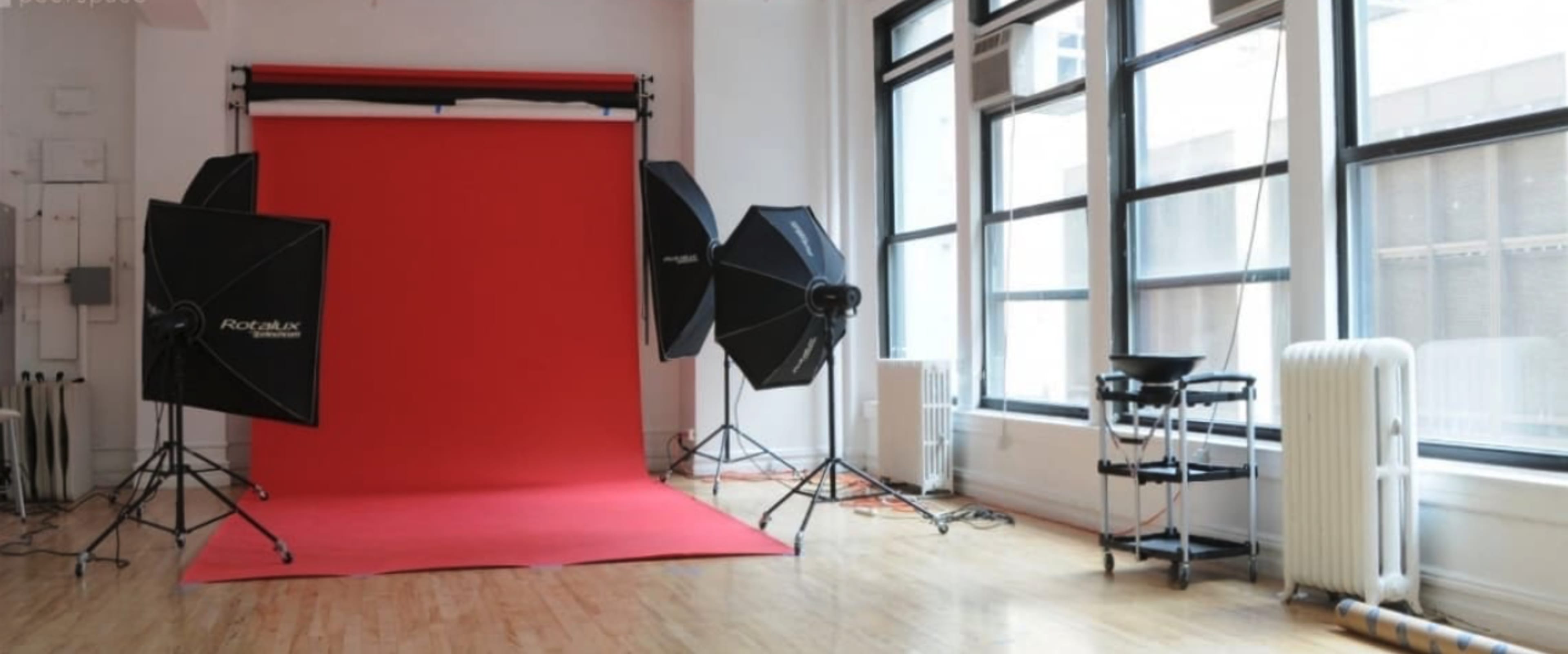 The image shows a photography studio with a red backdrop, softbox lighting, and large windows.