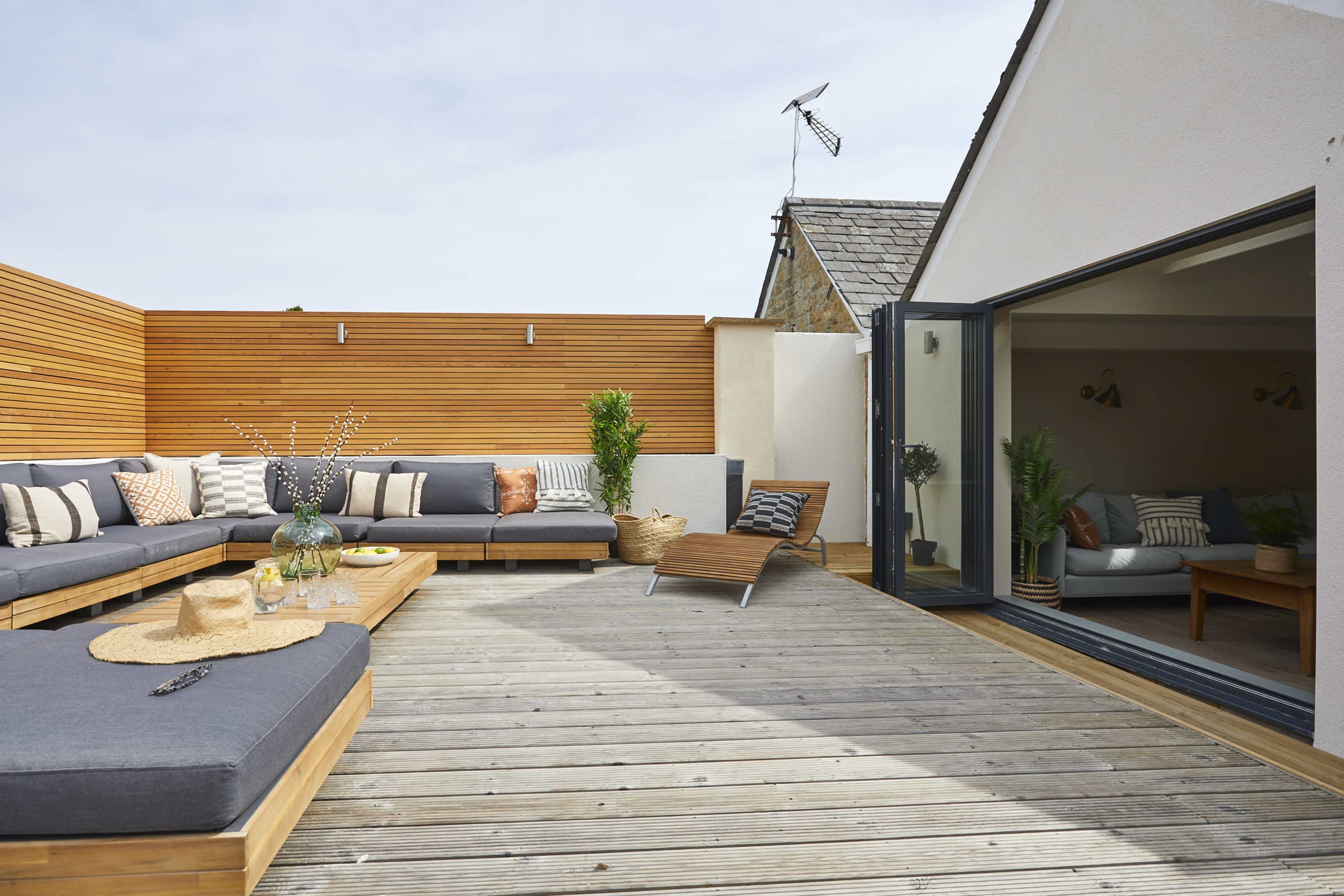 A modern outdoor lounge area features a wooden deck with sectional seating, a coffee table, and a sun lounger, surrounded by wooden privacy panels and potted plants.