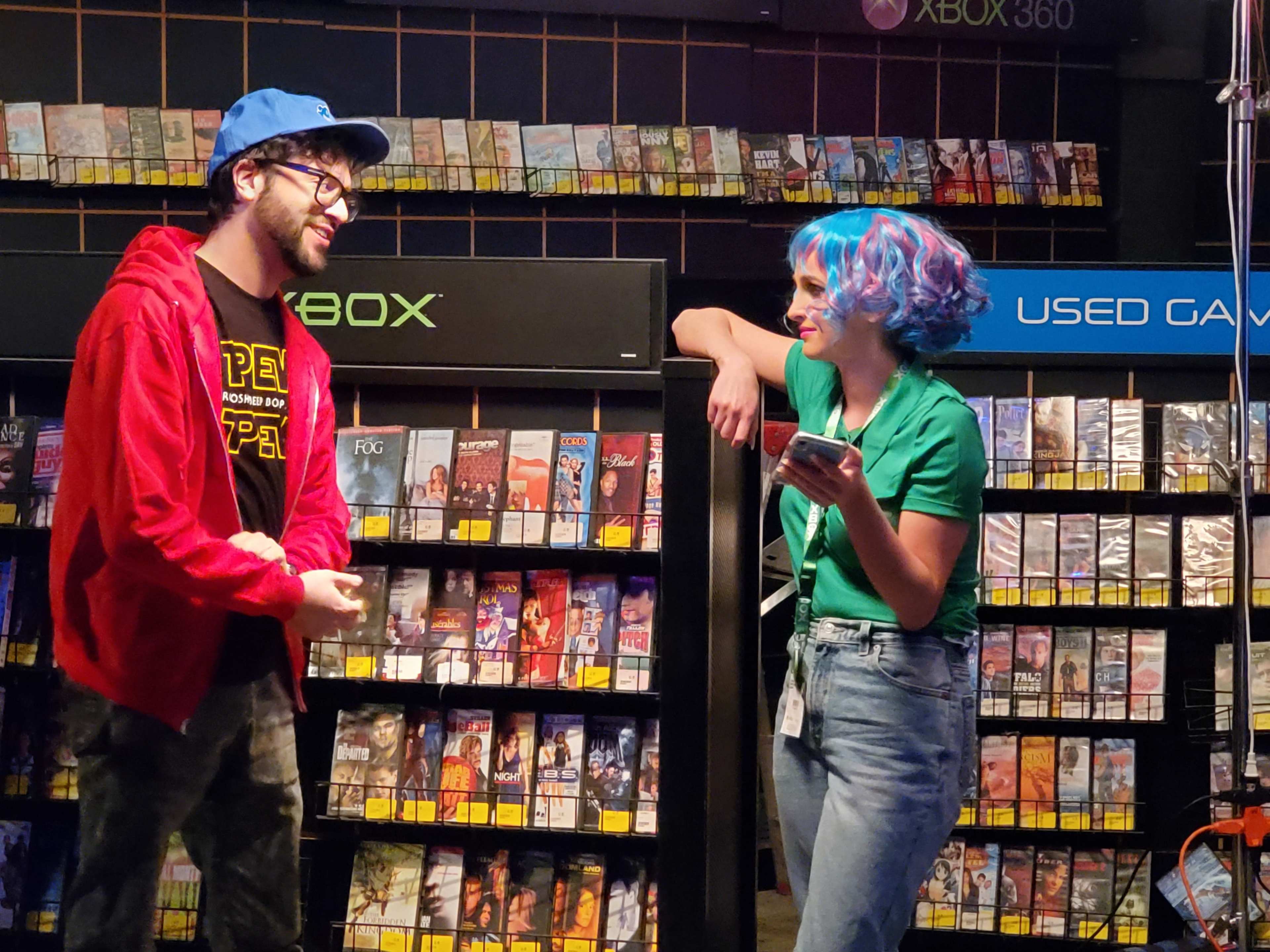 Two people engage in conversation in a retro video game store surrounded by shelves filled with games and consoles.