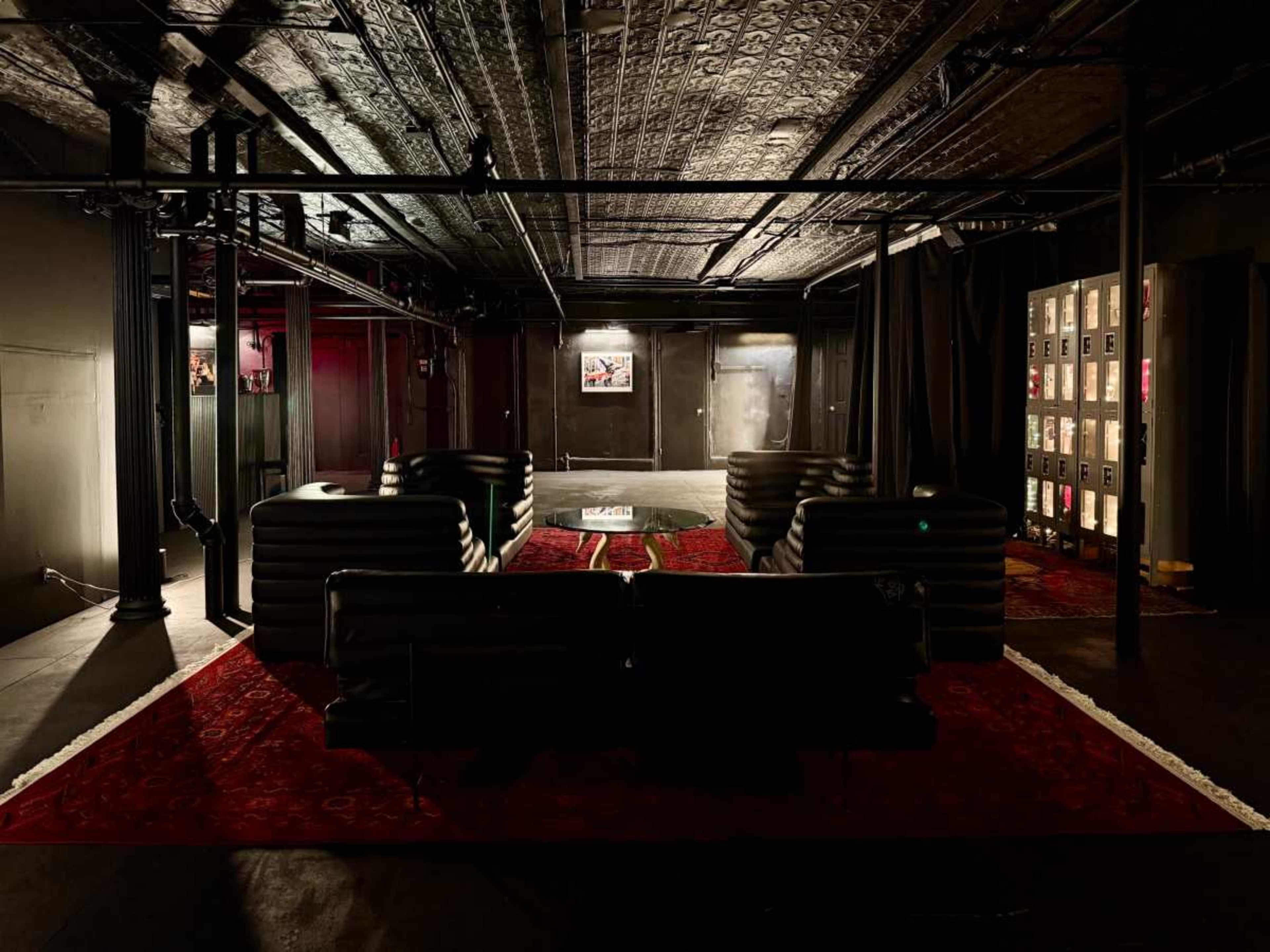 The image shows a dimly lit lounge area with black couches arranged around a table, set against a backdrop of exposed pipes and textured walls.