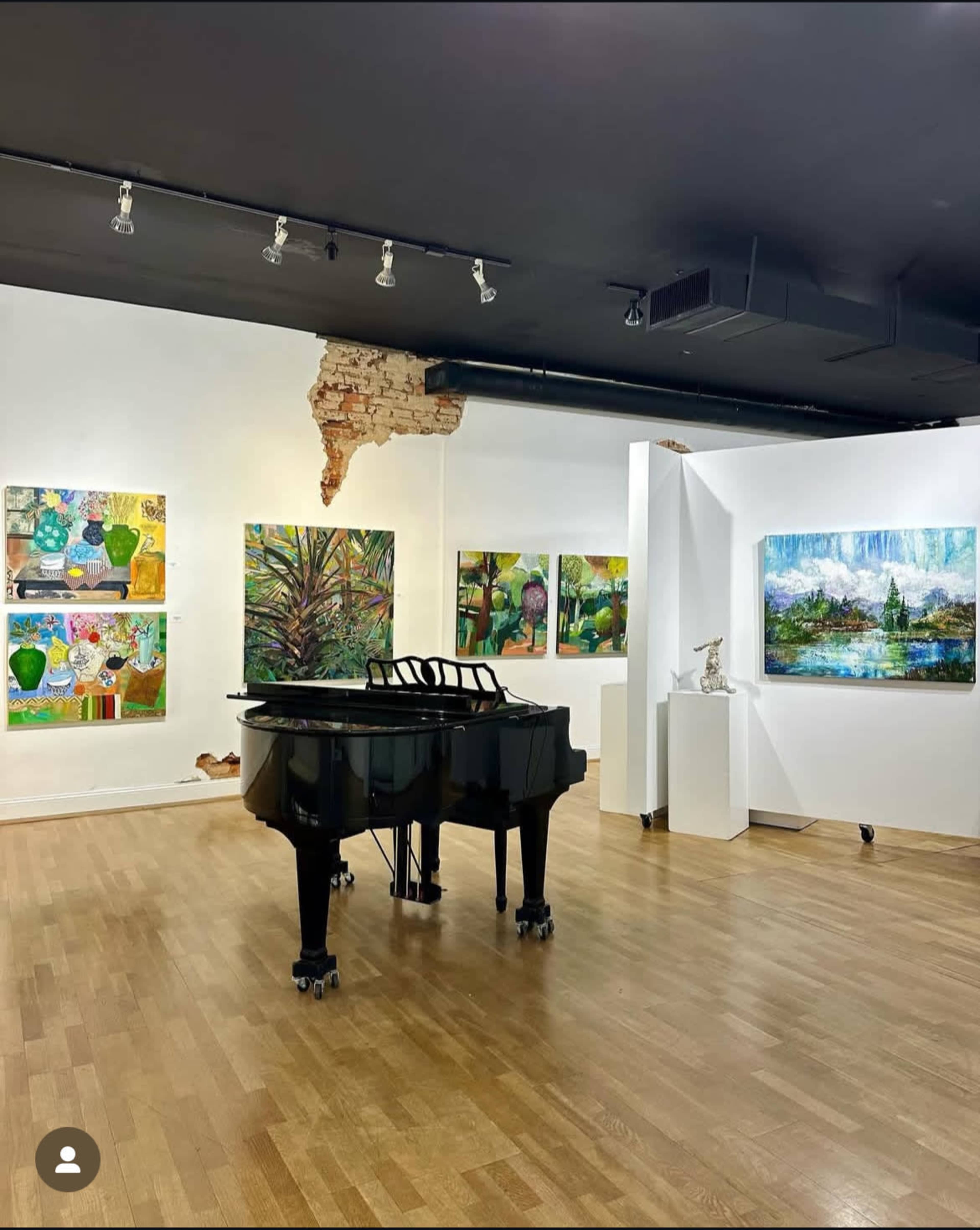 A grand piano occupies the center of an art gallery, surrounded by colorful paintings displayed on white walls.