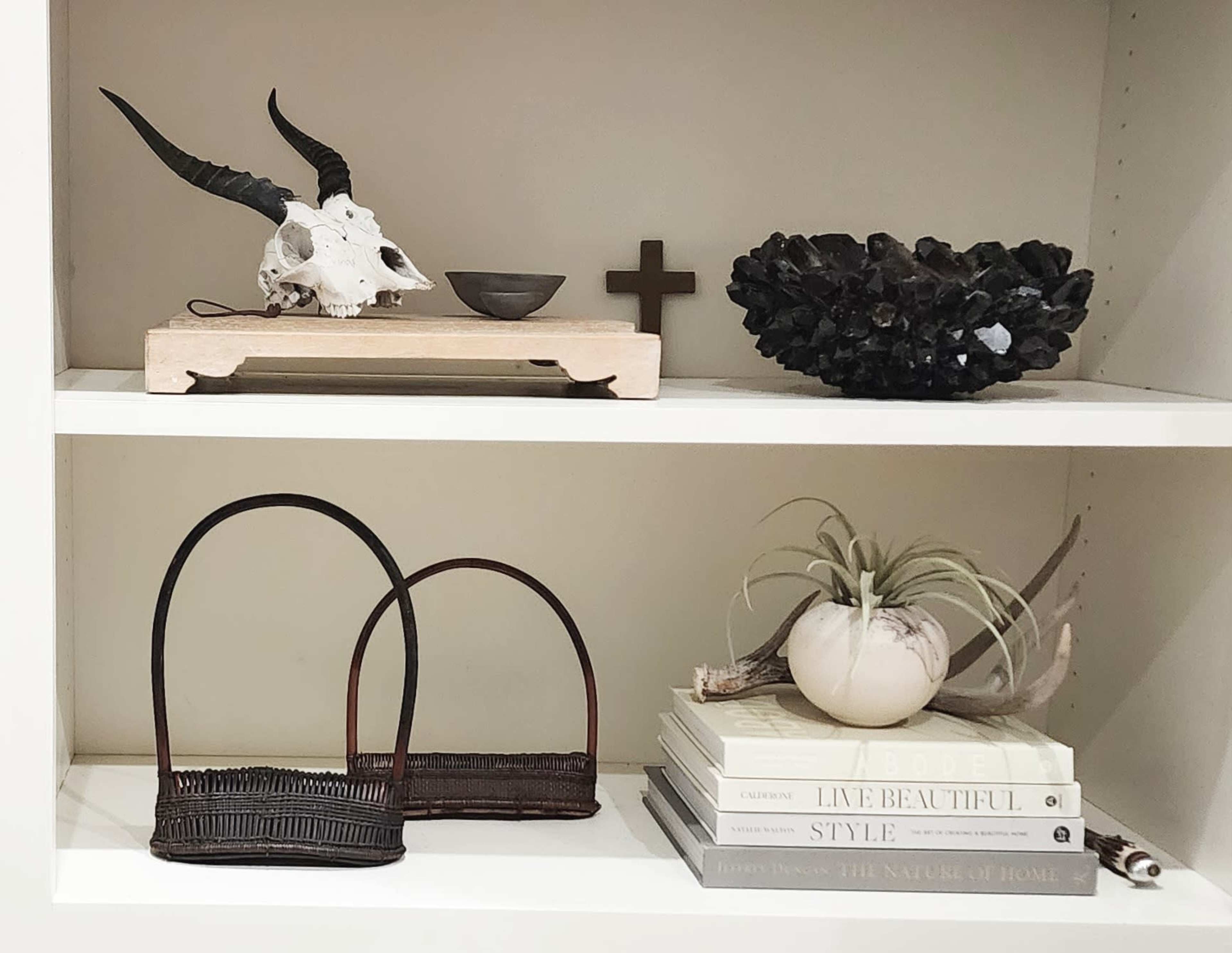 A shelf with decorative items, including a skull, a wooden bowl, a cross, a crystal cluster, woven baskets, books, and a plant.