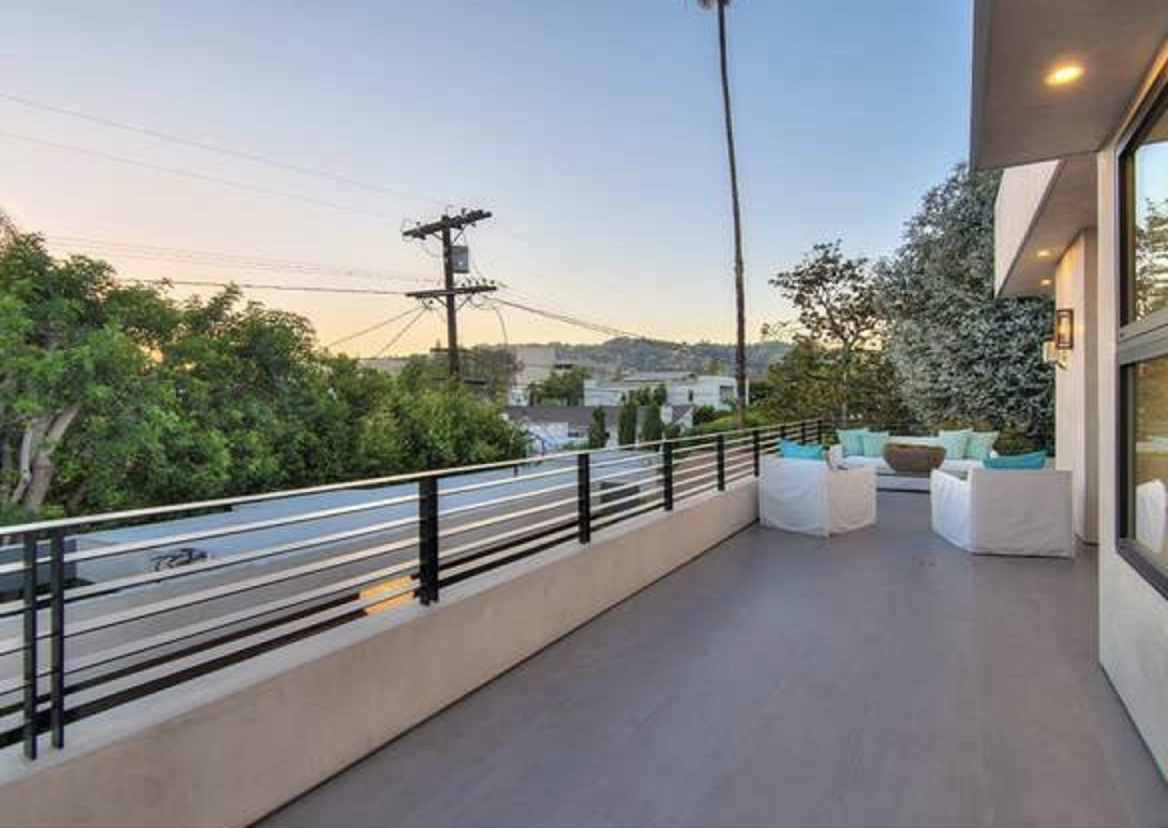 The image shows a modern balcony with minimalist seating, overlooking trees and utility poles against a clear sky.