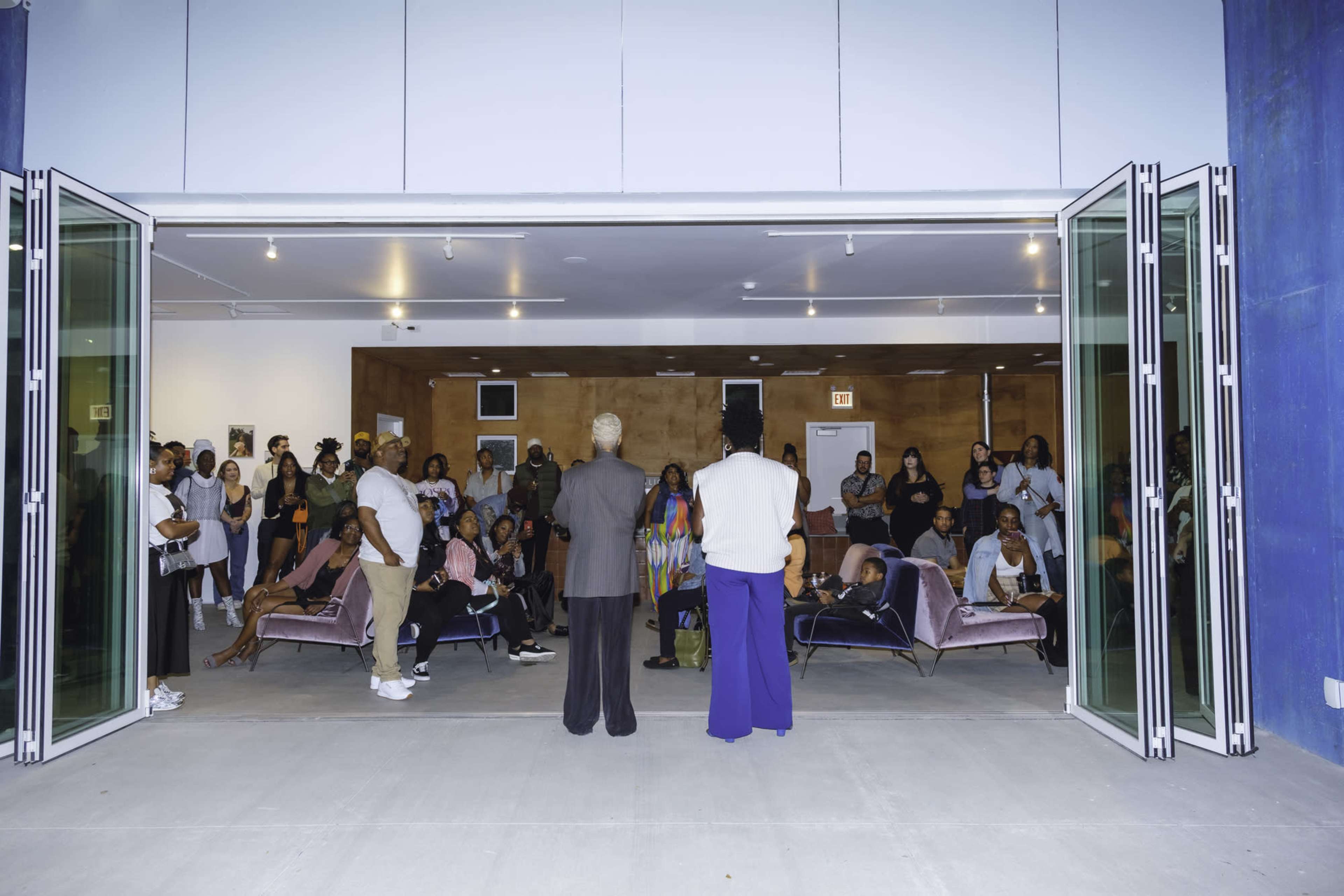 A group of people gathers inside a space with large open doors, seated and standing, while two individuals face the audience.
