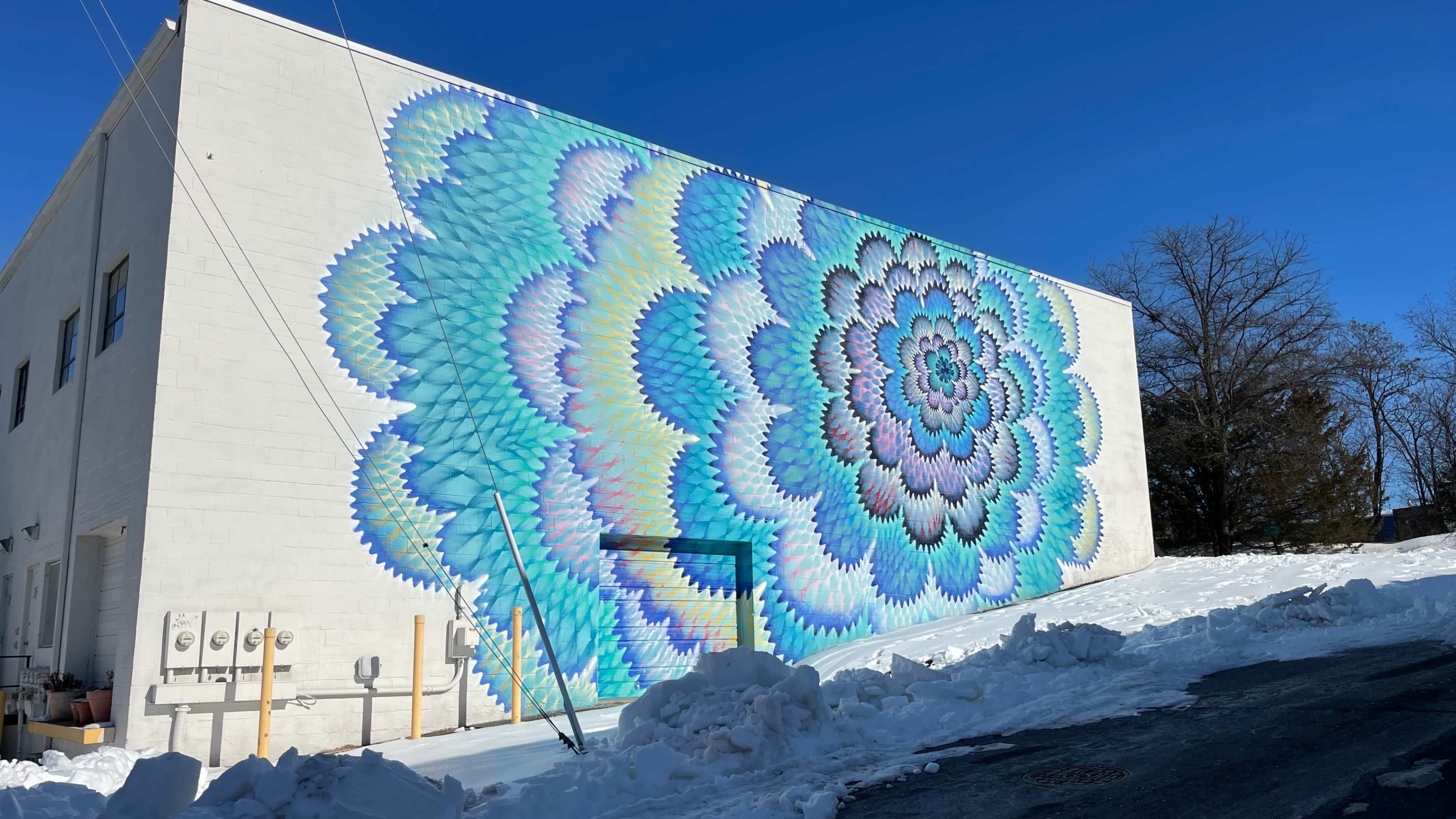 A large, colorful mural featuring a spiral design in shades of blue and green is painted on the side of a white building, with snow covering the ground in the foreground.