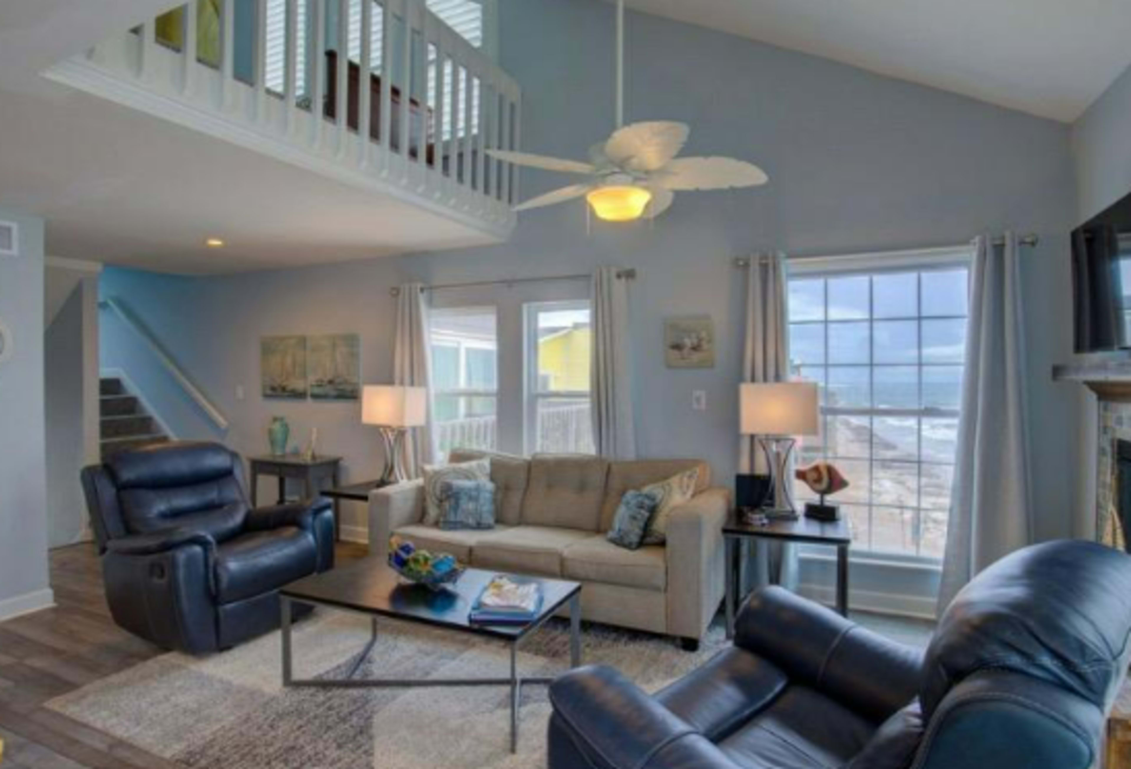 A bright living room with a sofa, two armchairs, and large windows facing a beach view, complemented by a lofted area above.