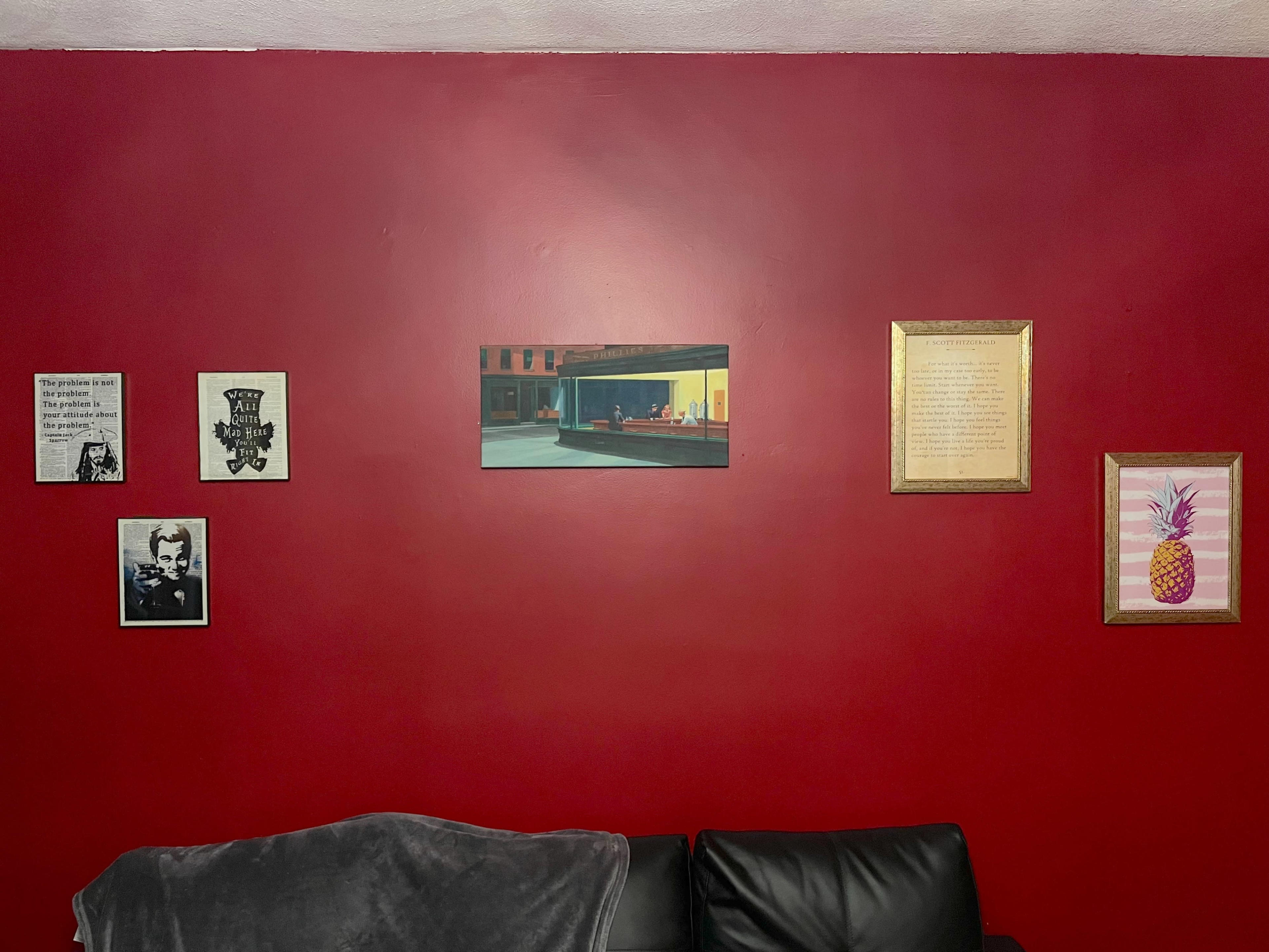 A red wall features a variety of framed artworks and posters, including a painting of diners and images of notable figures, above a black sofa with a grey throw blanket.