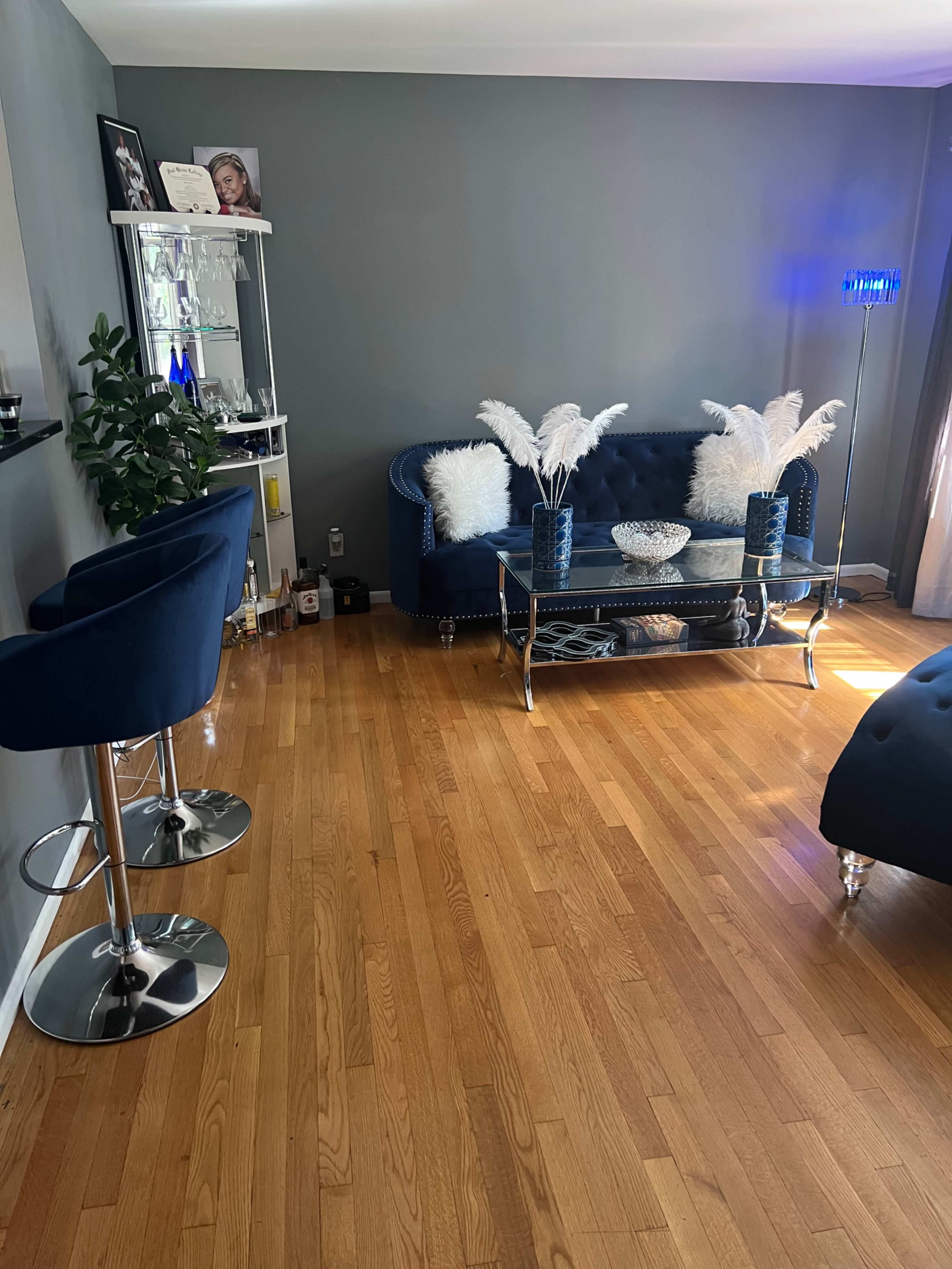 A living room featuring a blue sofa, a glass coffee table with decorative items, and two bar stools on a wooden floor.