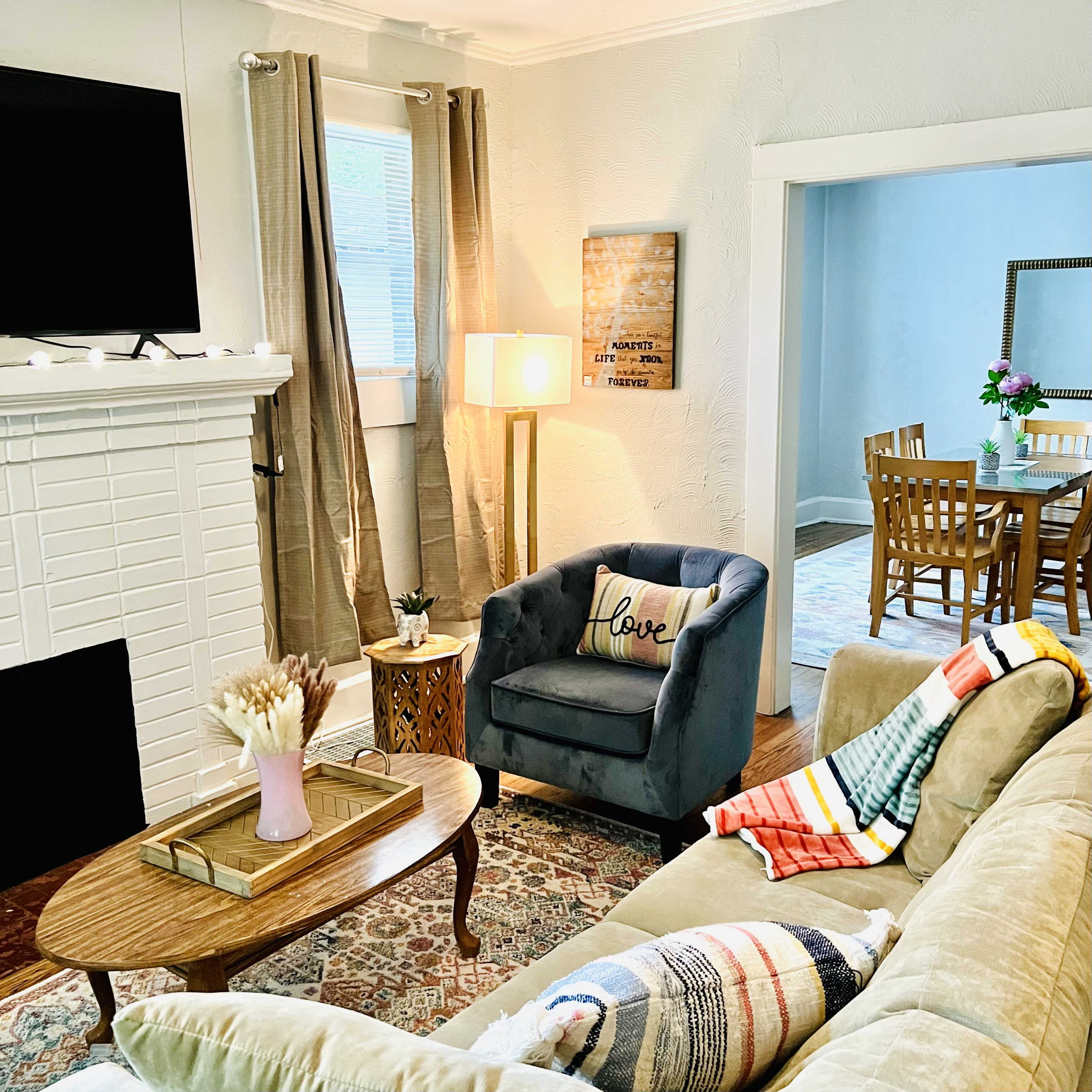 The living room features a blue armchair with a "love" cushion, a wooden coffee table, and a fireplace, with a dining area visible in the background.