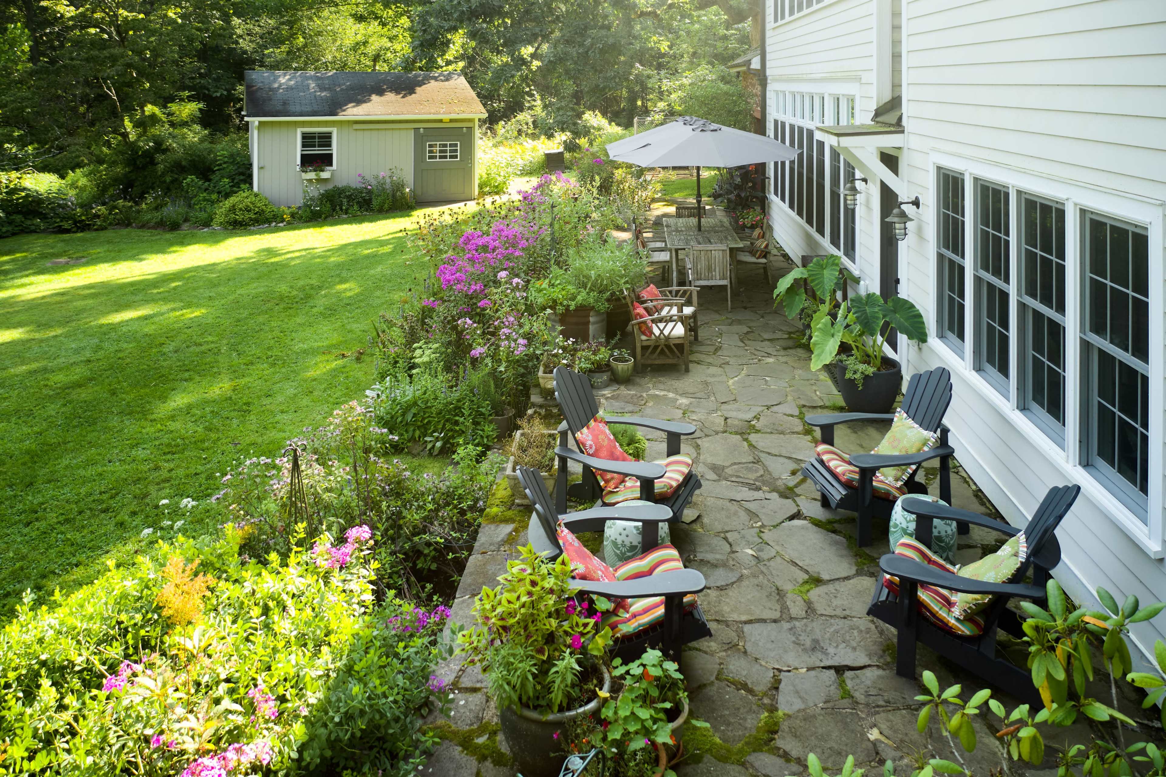 A stone patio with four wooden chairs and colorful cushions overlooks a garden filled with flowers and greenery, alongside a white house and a shed.