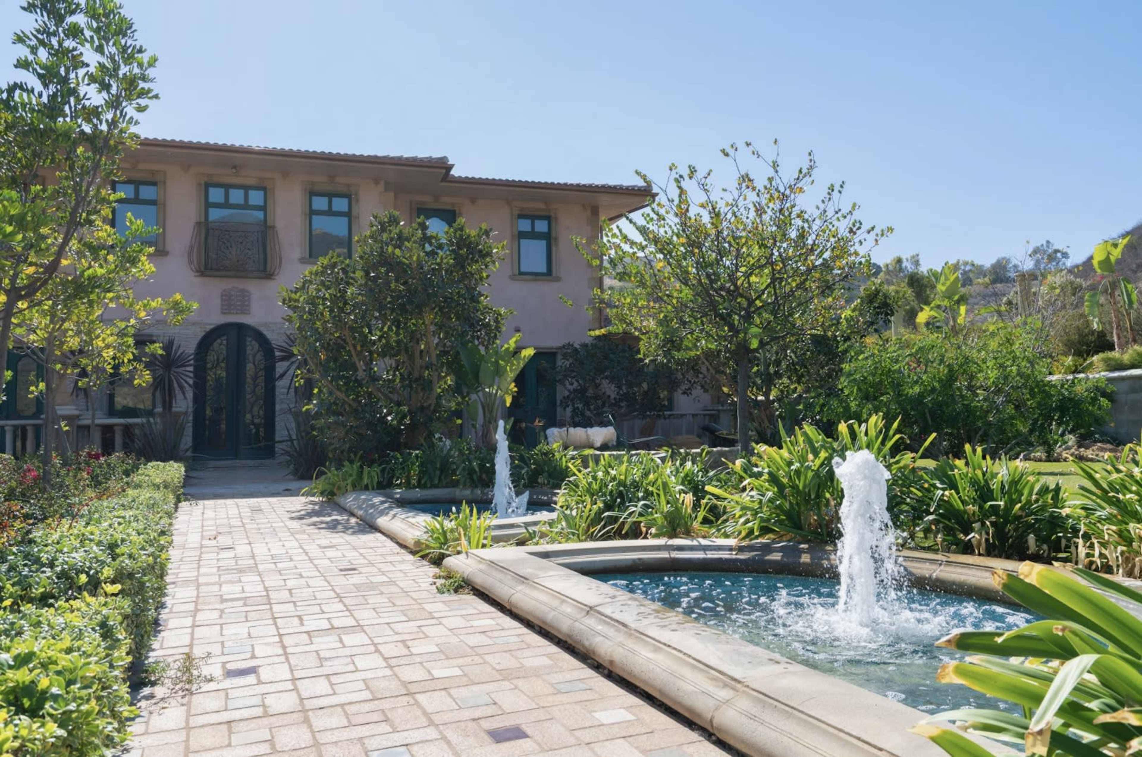 A landscaped pathway leading to a two-story house, featuring a fountain and surrounding greenery.