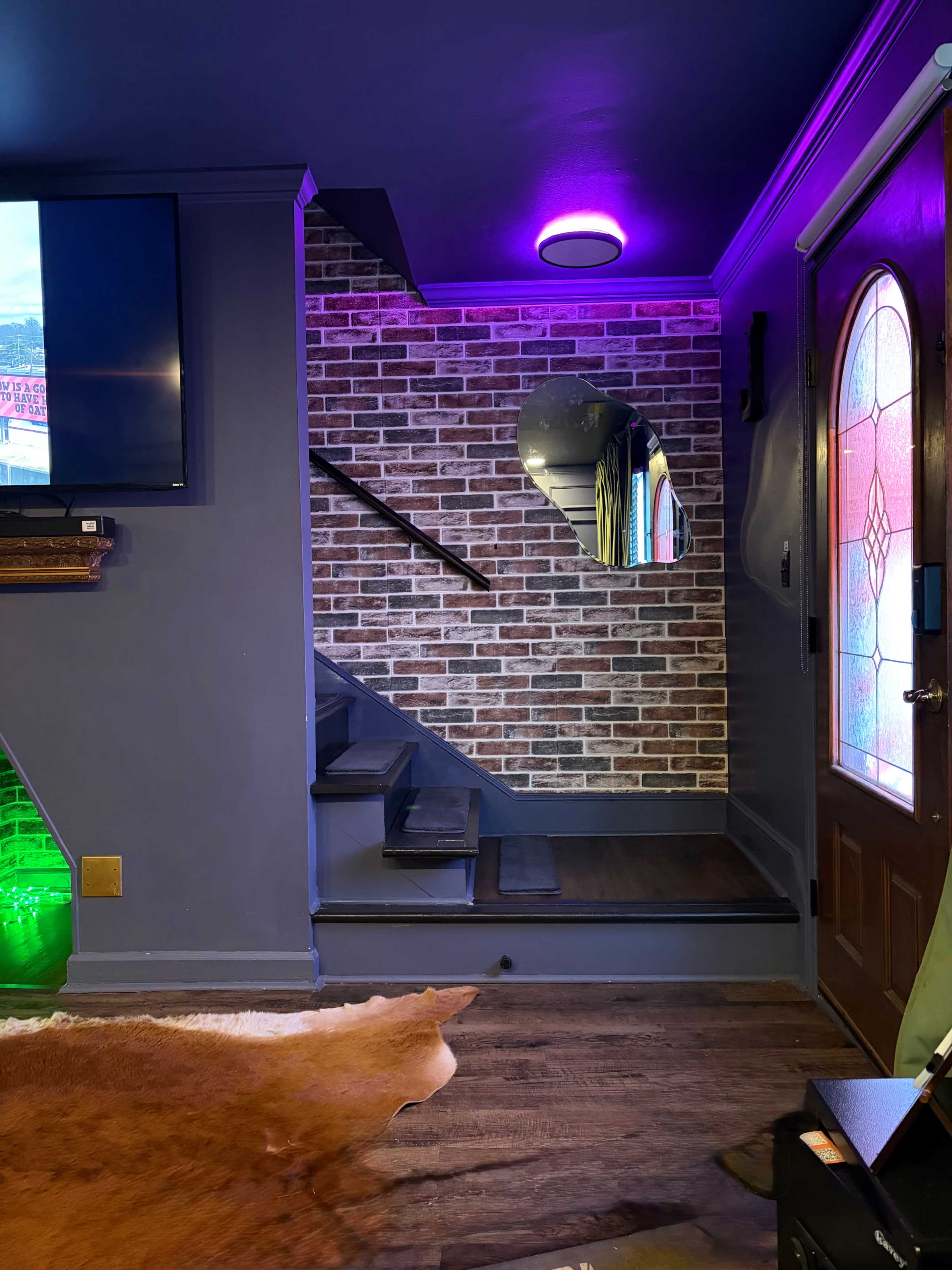 The image shows a modern entryway featuring a staircase with a brick accent wall, a round mirror, a television, and colorful lighting.