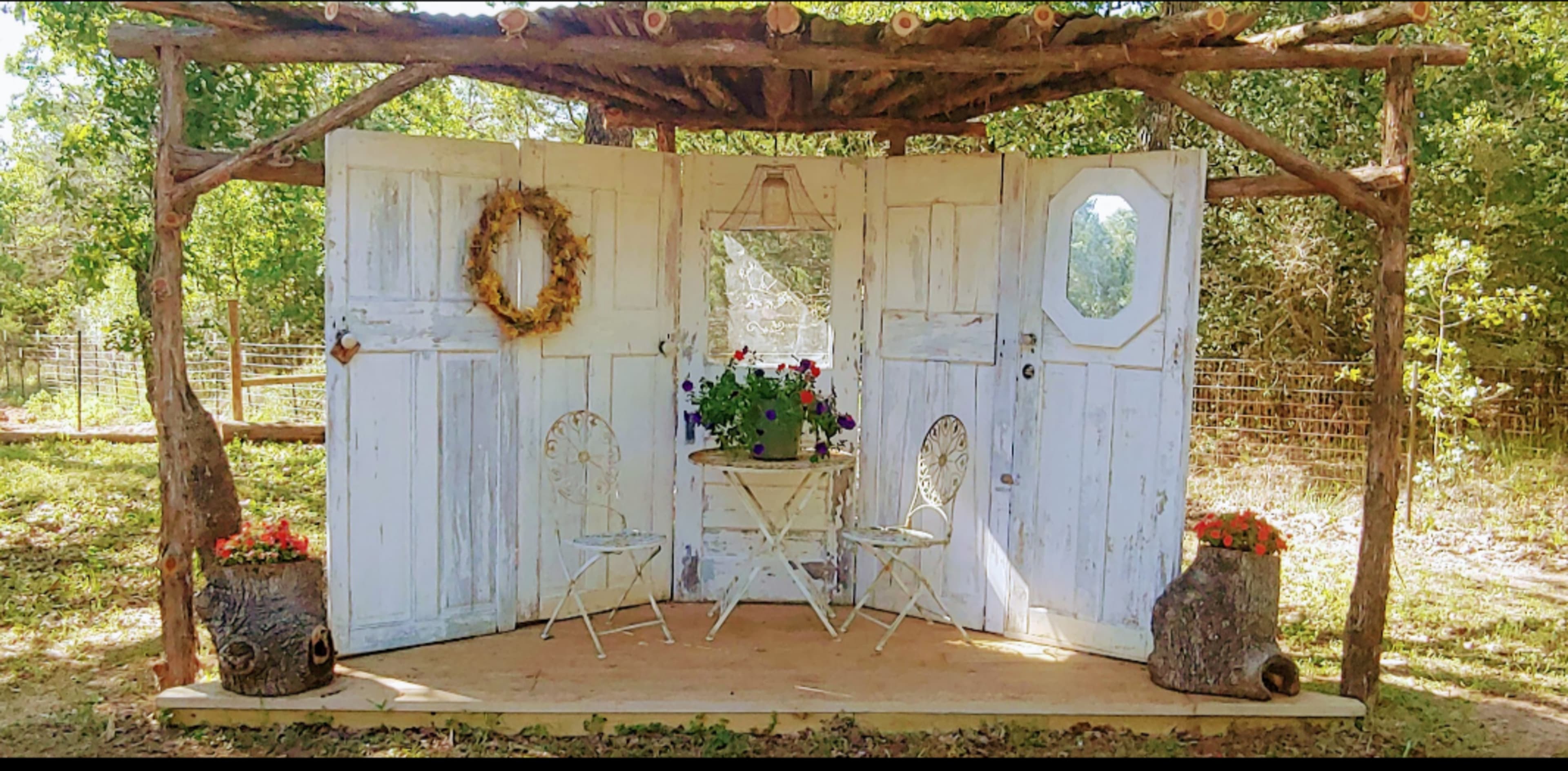 The image shows an outdoor setting featuring a rustic wooden structure with multiple white doors, a small round table with a floral arrangement, and two chairs.