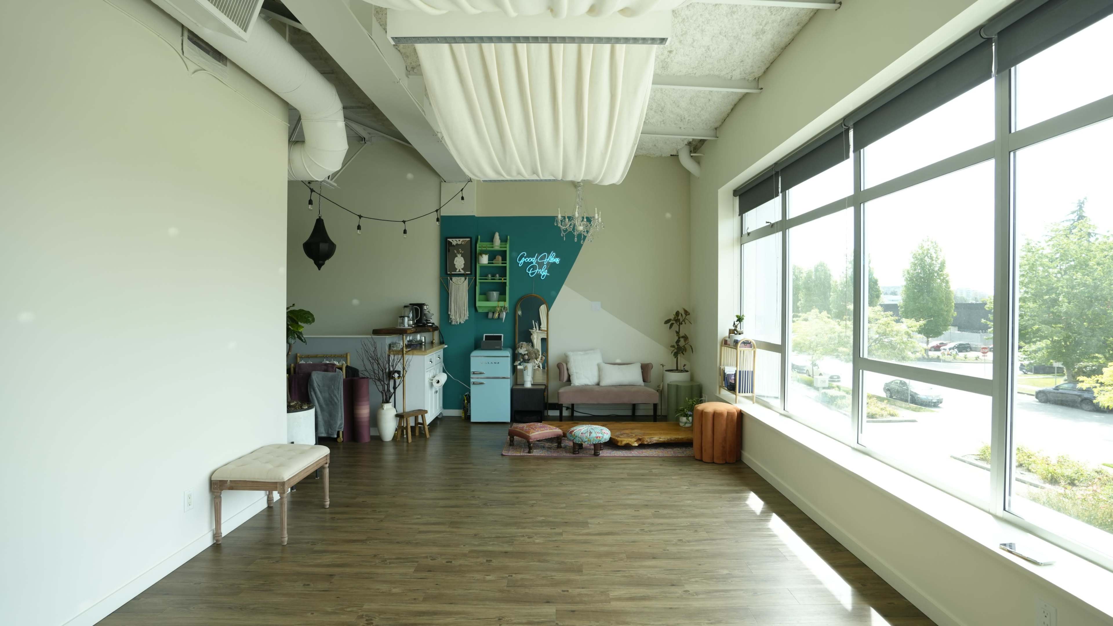 The image shows a spacious, well-lit interior with large windows, featuring a seating area, decorative plants, and a small kitchenette in the background.