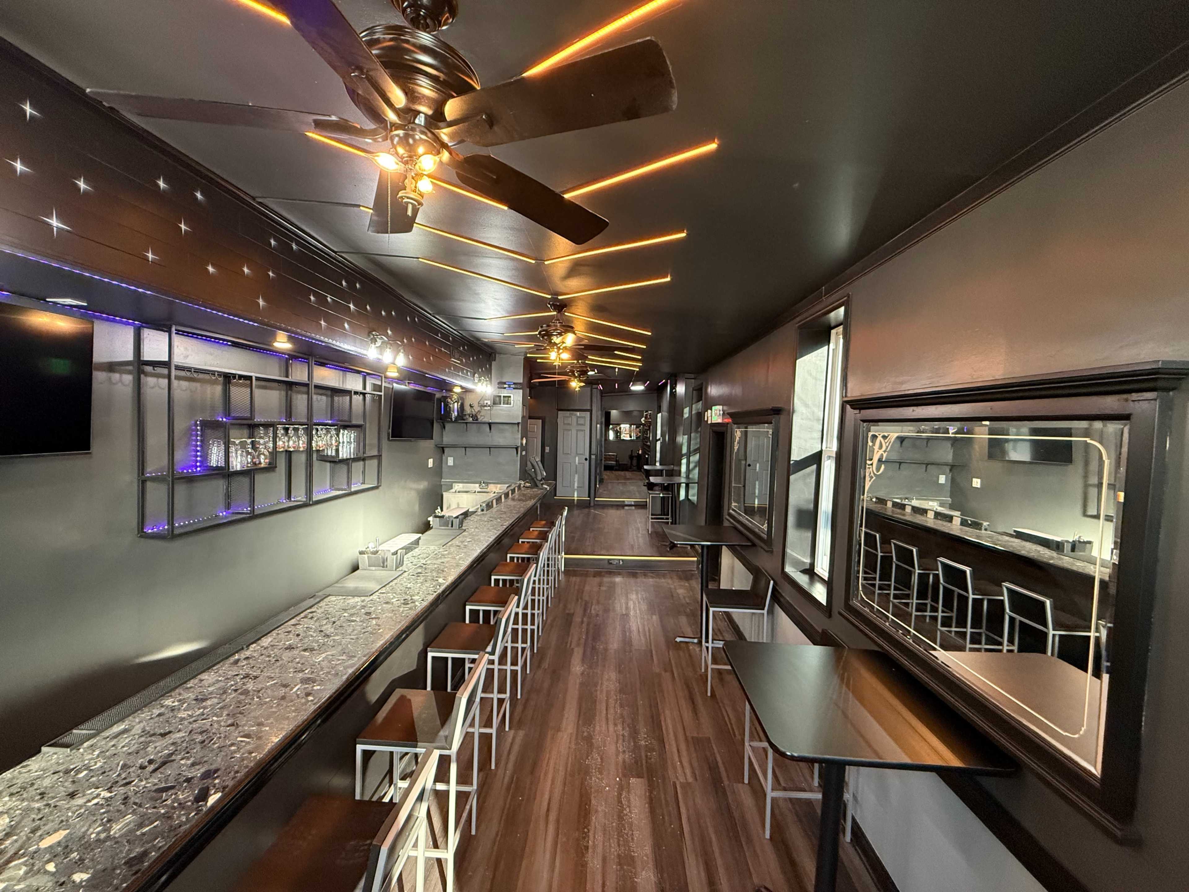 The image shows a modern café or bar interior featuring a long, gray granite countertop, white bar stools, and warm lighting against dark walls, with a few tables set up along the side.