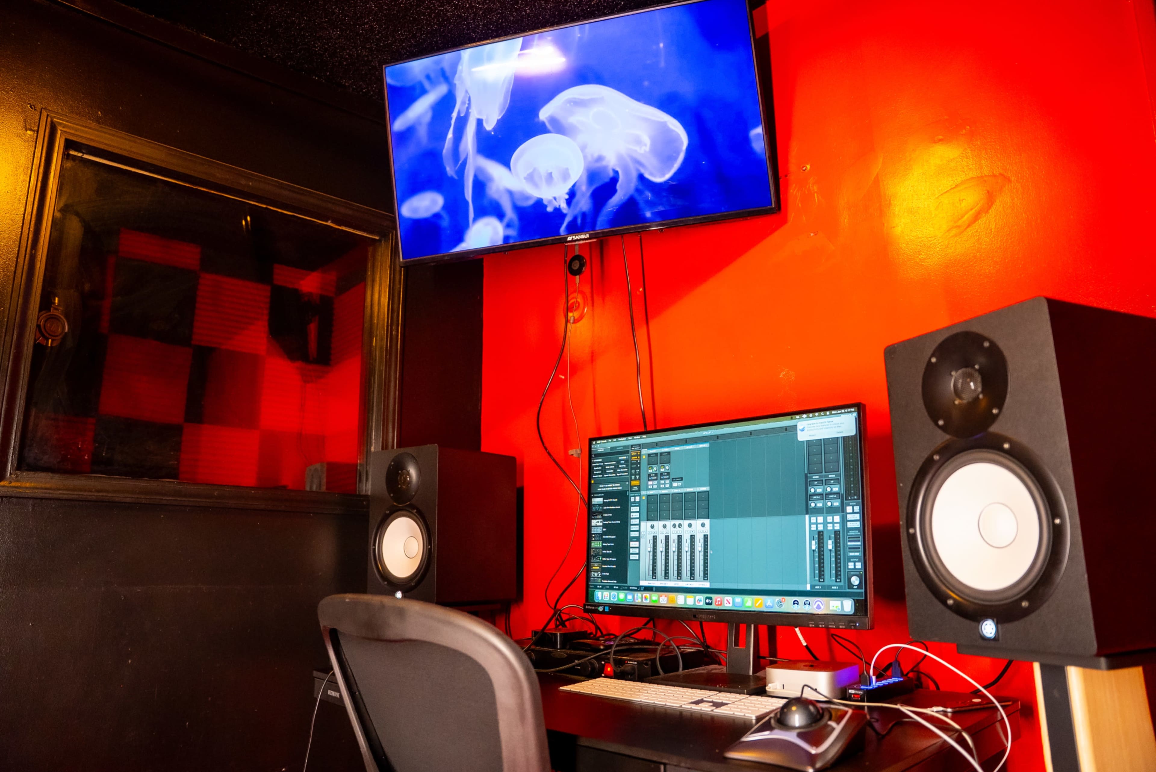 The image shows a recording studio with a computer, audio monitors, and a television displaying jellyfish, set against a red wall.