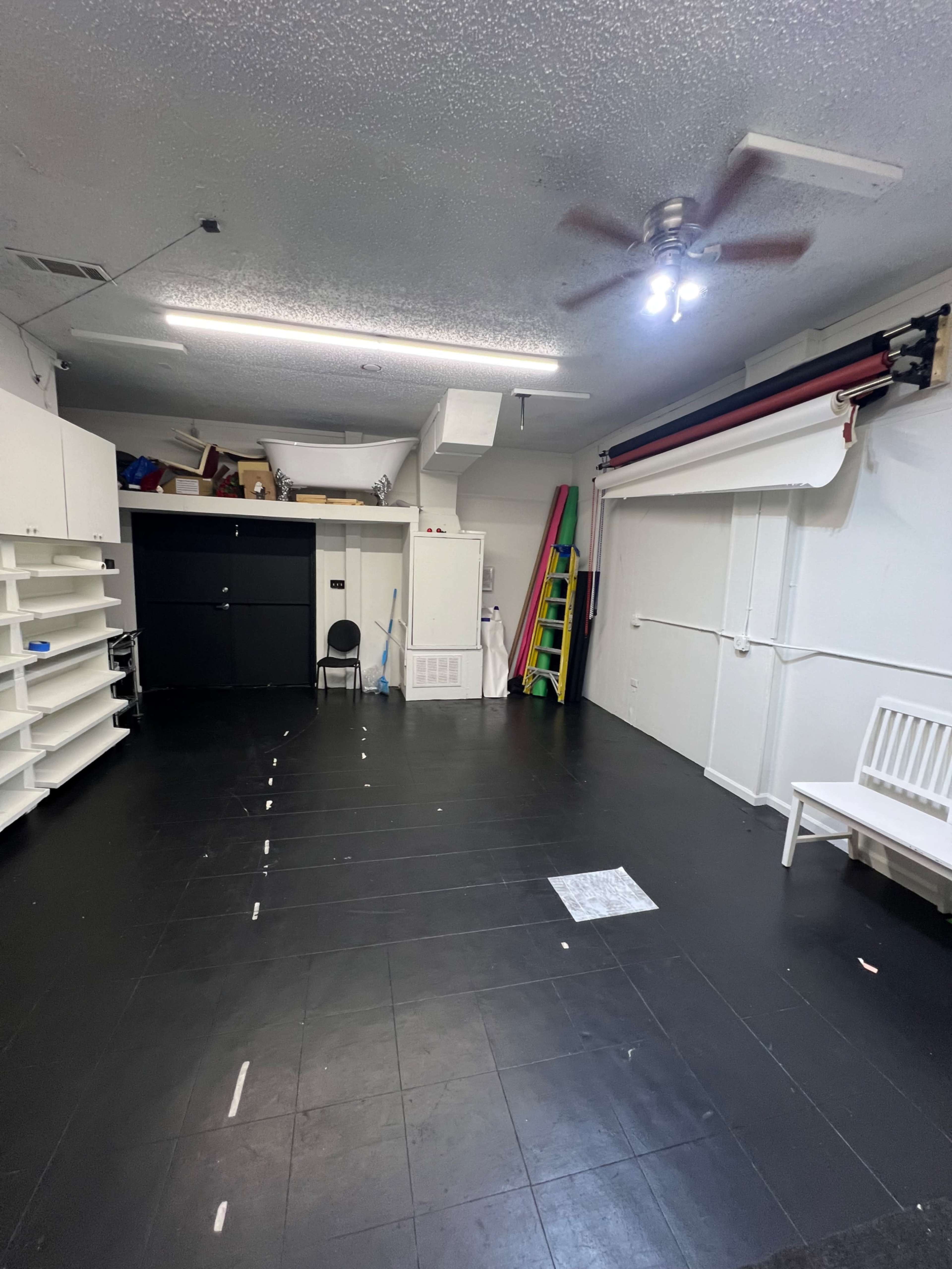 A sparsely furnished room with black tiled flooring, white shelving units on the left, a chair, and a large roll of backdrop material along the right wall.