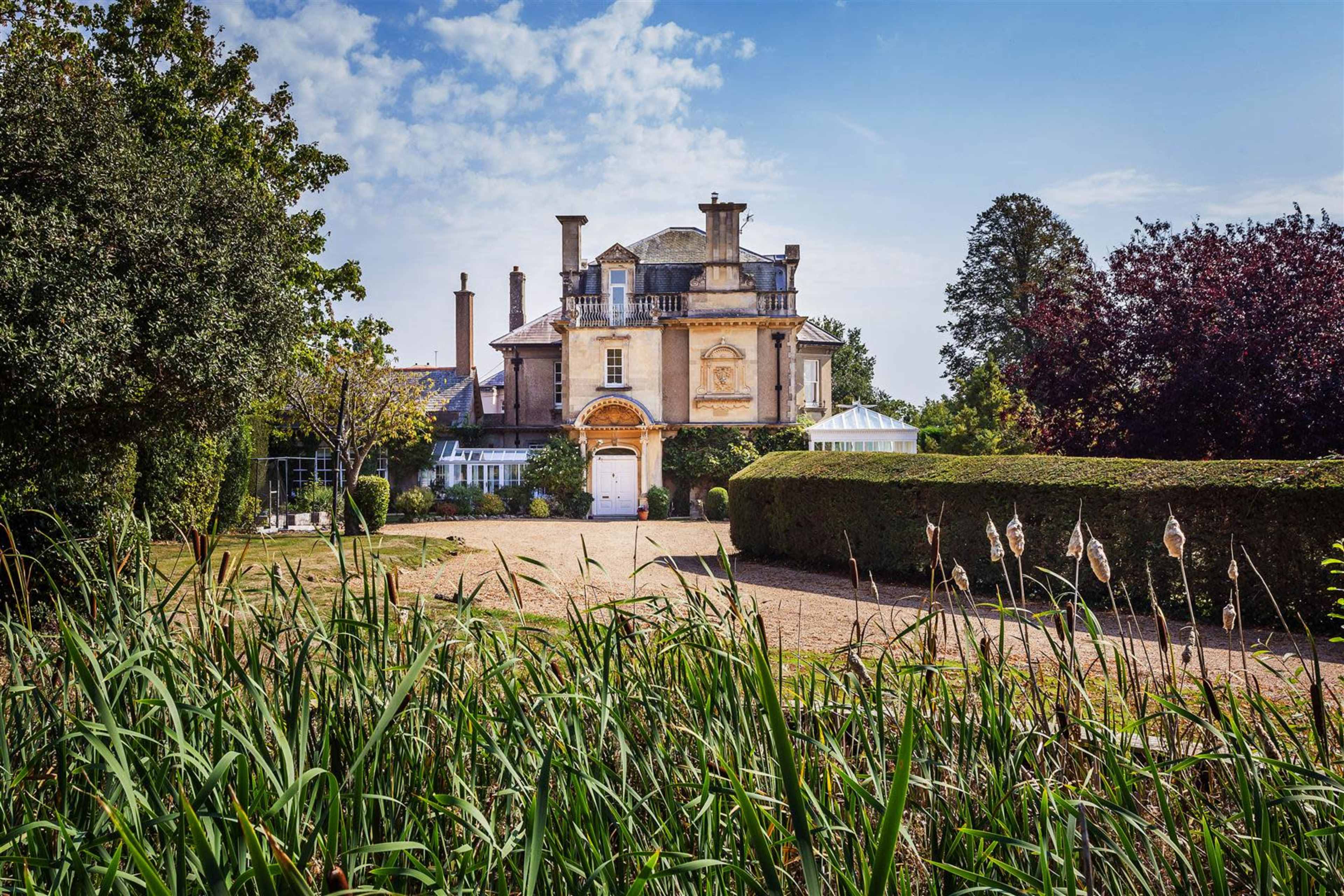 A large, two-story mansion with ornate architectural features is surrounded by well-manicured gardens and tall grasses.