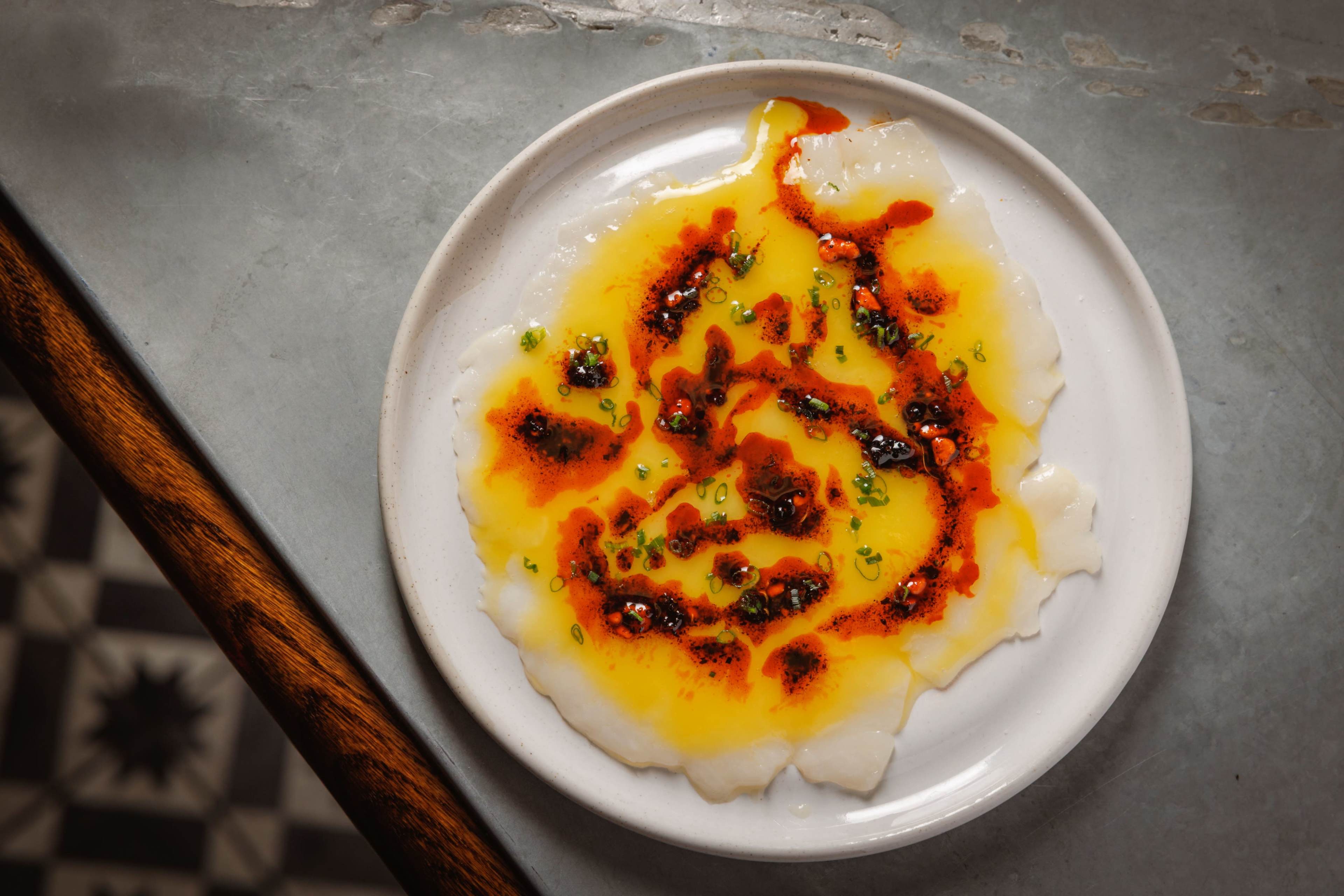 A white plate holds a dish of thinly sliced fish drizzled with a vibrant red and yellow oil and garnished with green herbs.