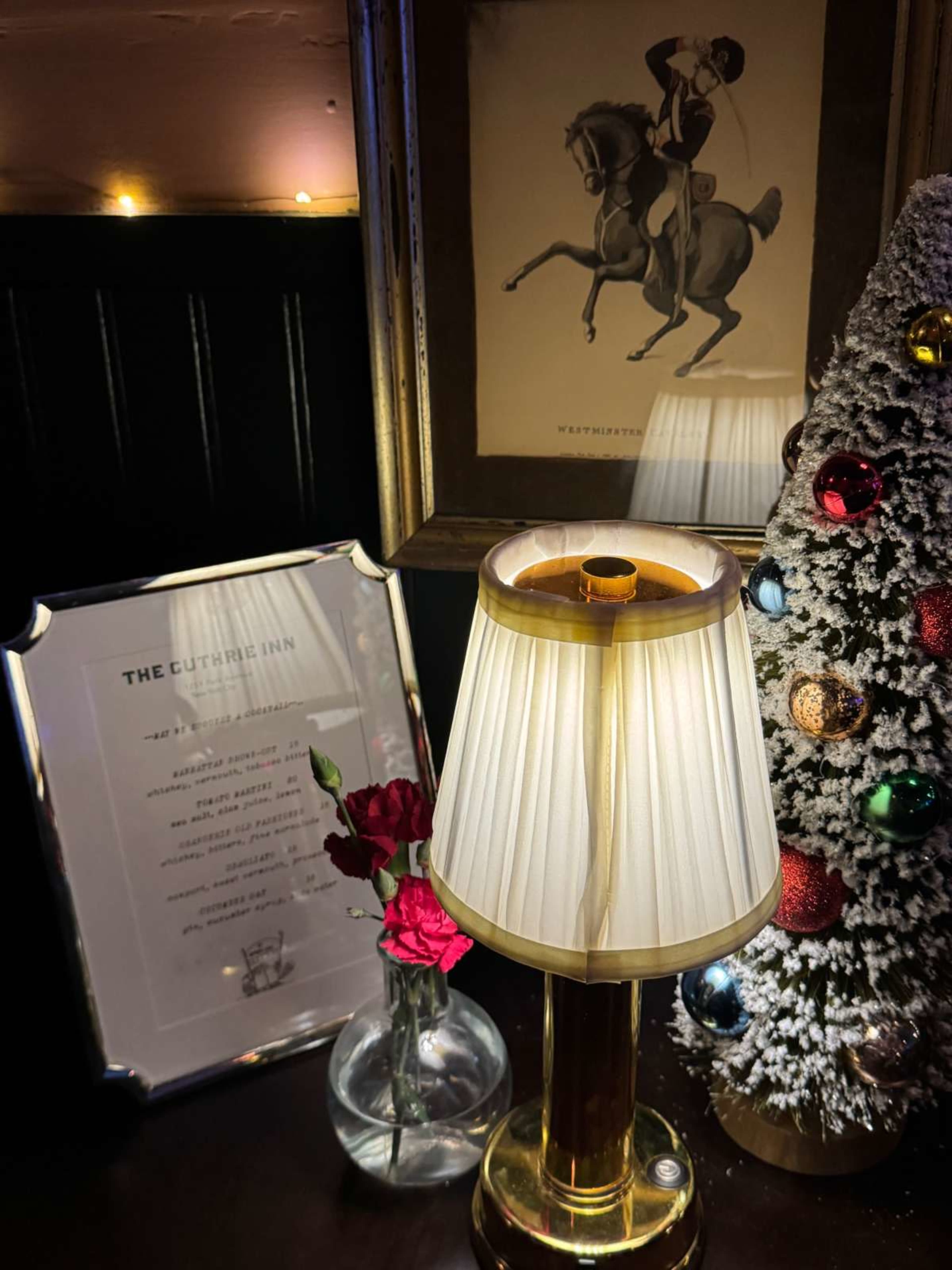 A decorative table features a lamp, a small vase with roses, a framed menu, and a Christmas tree adorned with ornaments.