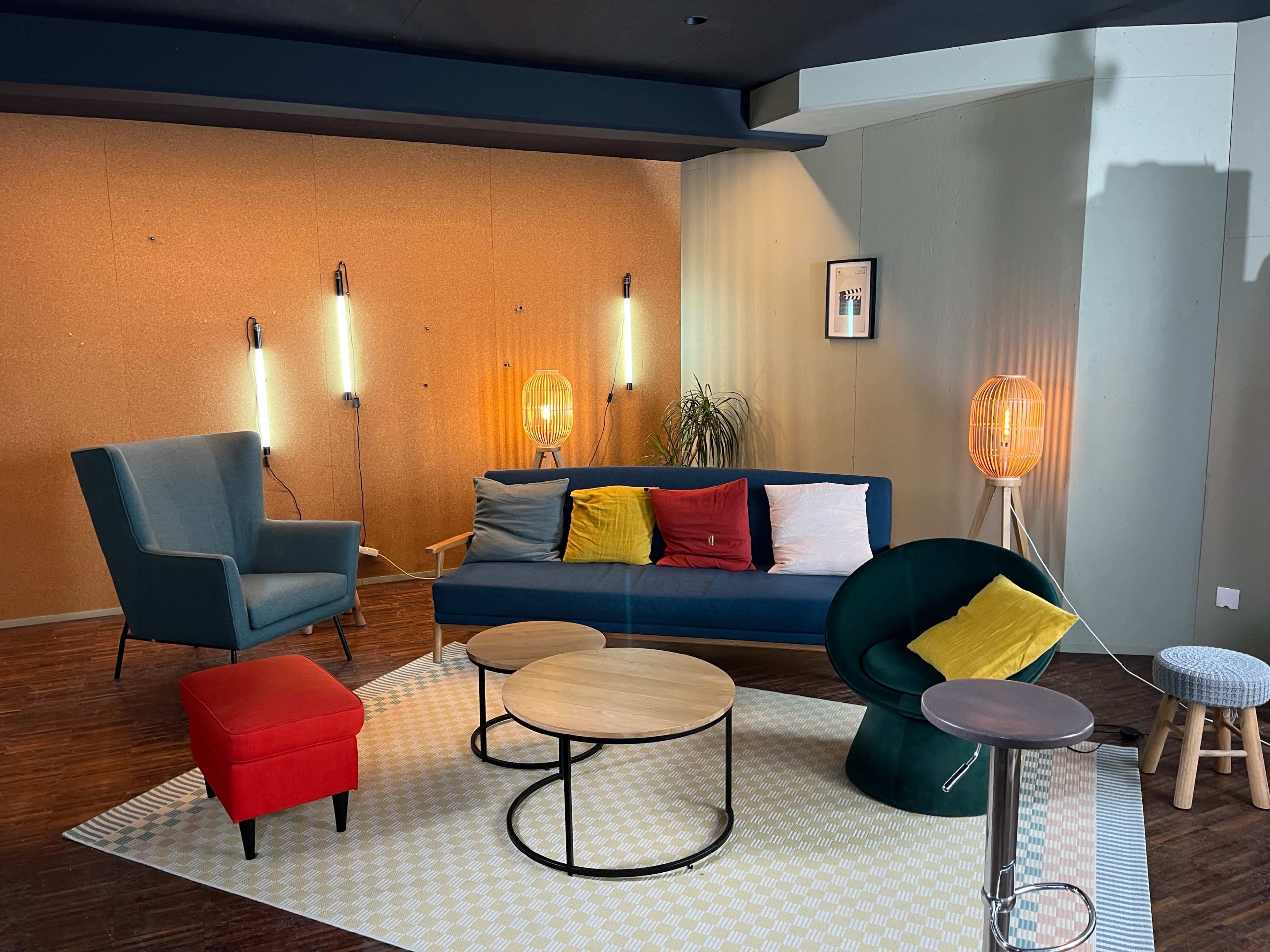 The image shows a modern living space featuring a blue sofa with colorful cushions, a round coffee table, and varied seating, complemented by unique lighting fixtures on the walls.