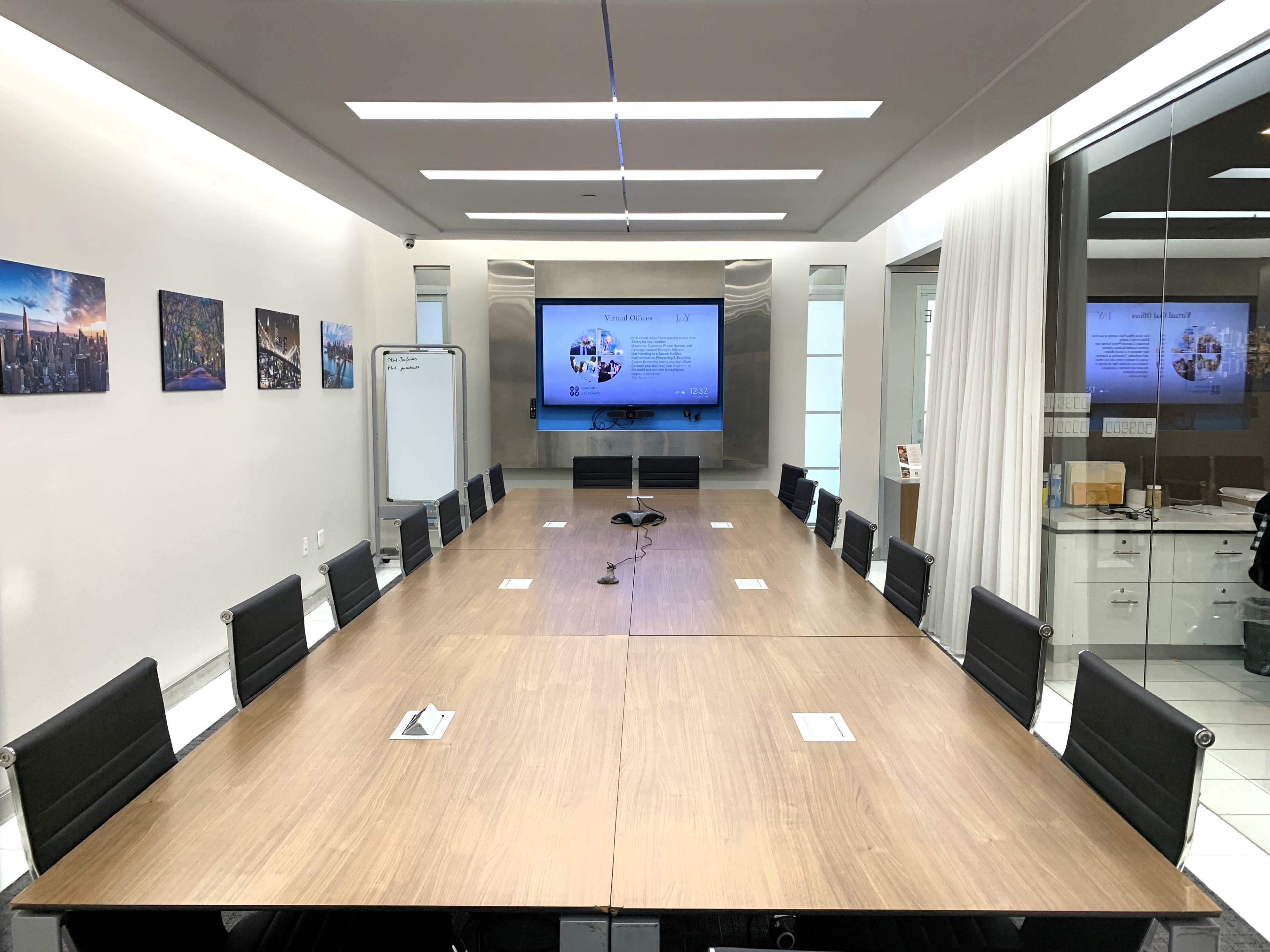 The image shows a modern conference room with a large wooden table, black chairs, a screen displaying information, and wall-mounted photos.