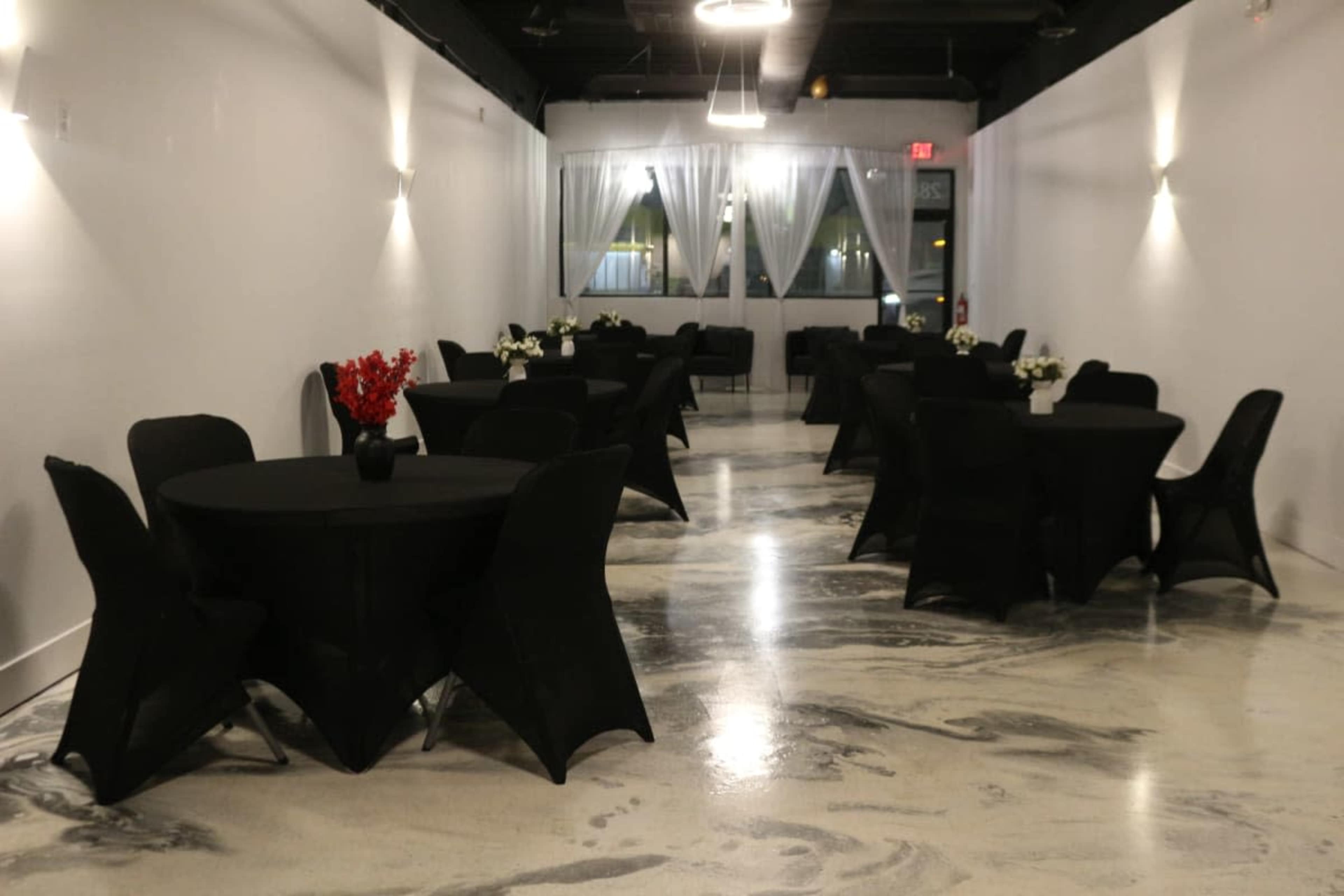 A spacious event room with round tables covered in black cloth, organized in a neat arrangement against a white wall backdrop.