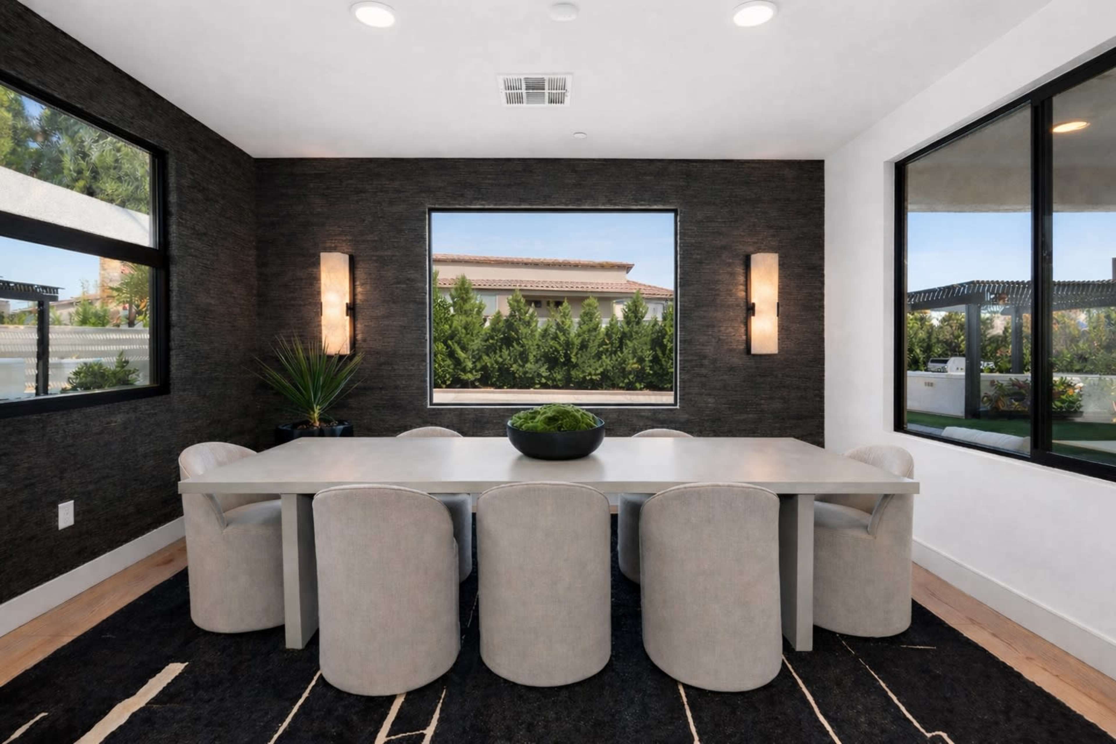 A modern dining room features a large, rectangular table surrounded by upholstered chairs, with large windows showcasing a view of greenery outside.