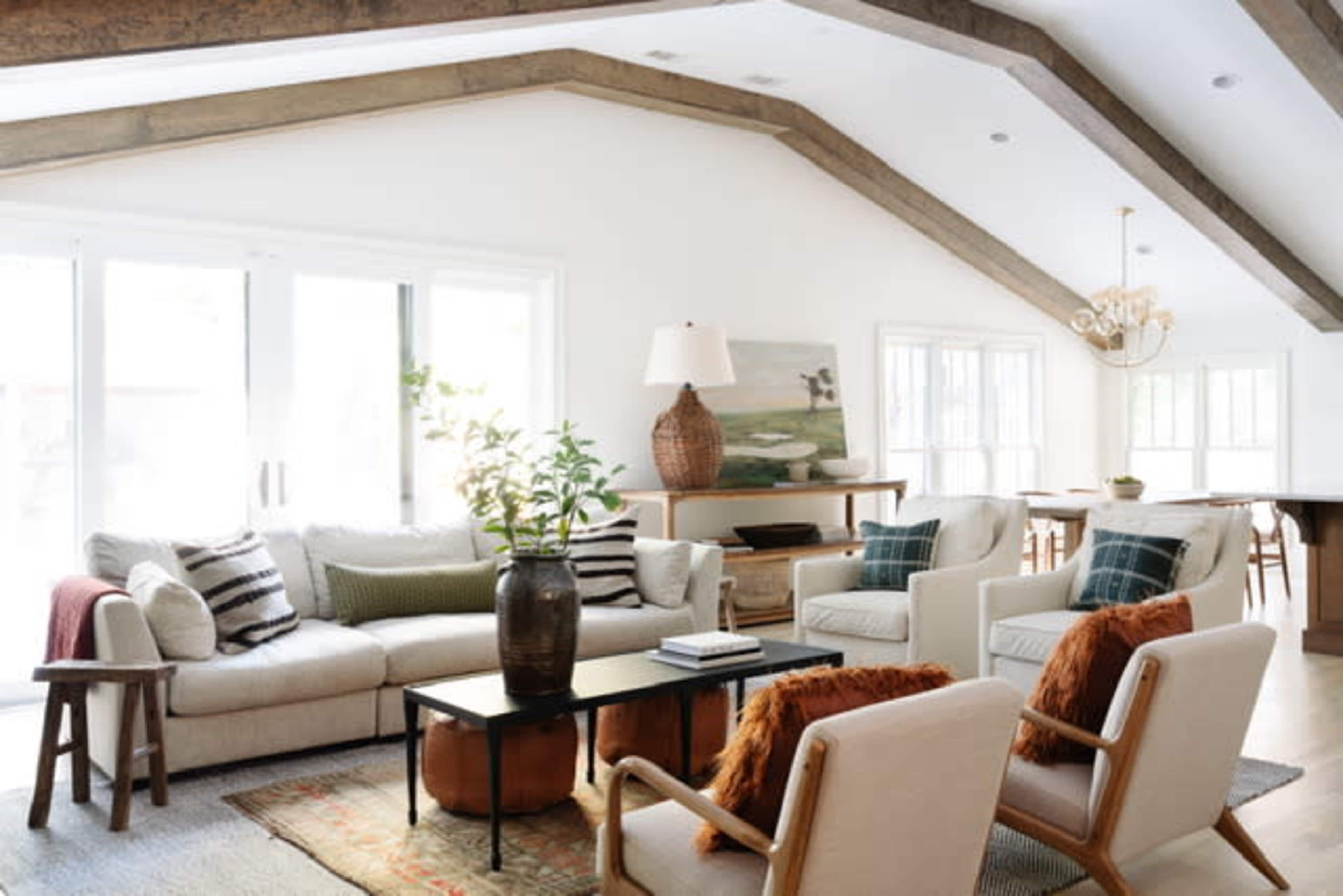 The spacious living room features a light-colored sofa, two armchairs, a rectangular coffee table, and large windows with wooden beams overhead.