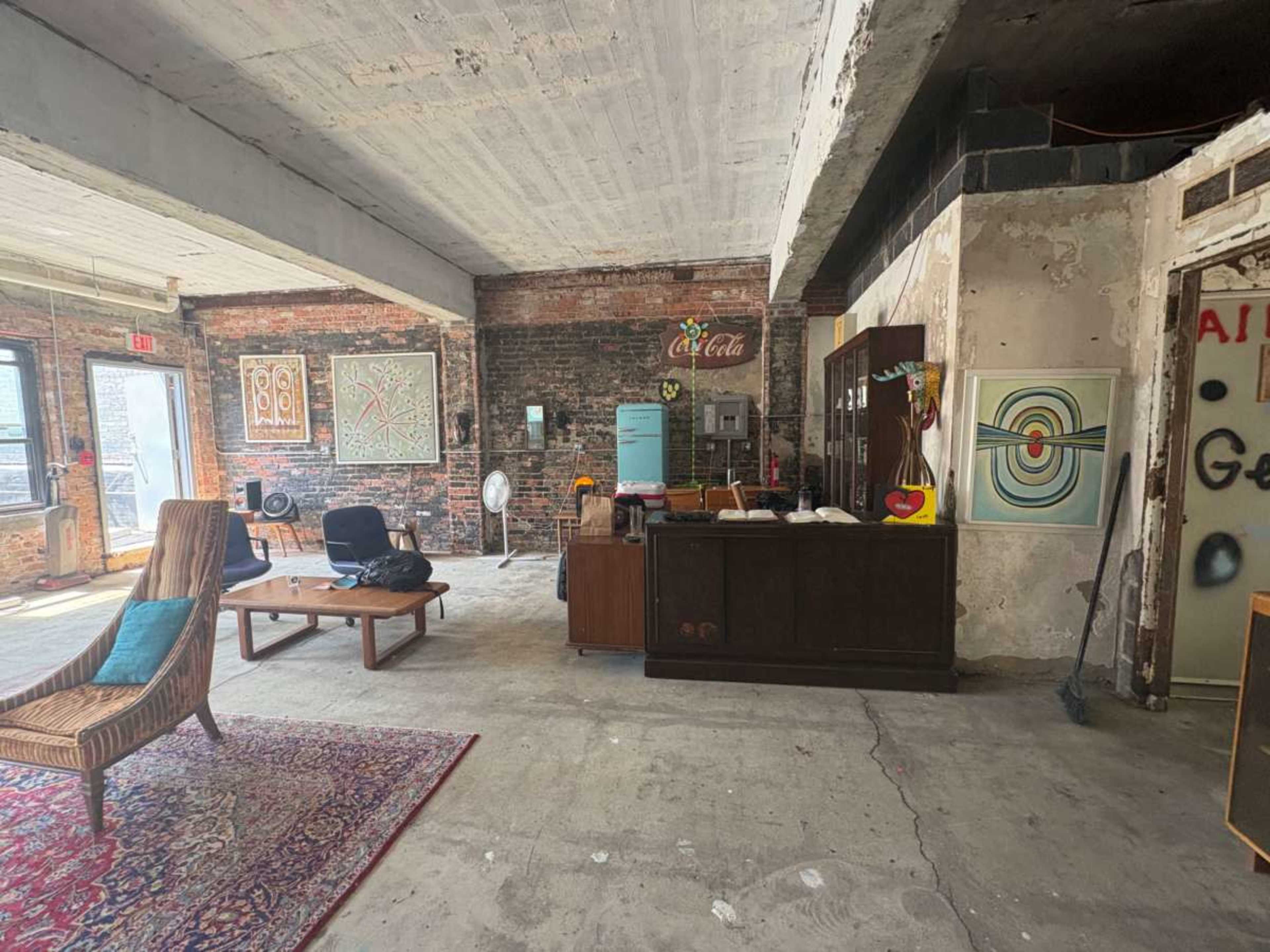 The image shows a spacious room with exposed brick walls, featuring a seating area with a chair and a coffee table, along with a vintage sideboard and various artworks on display.