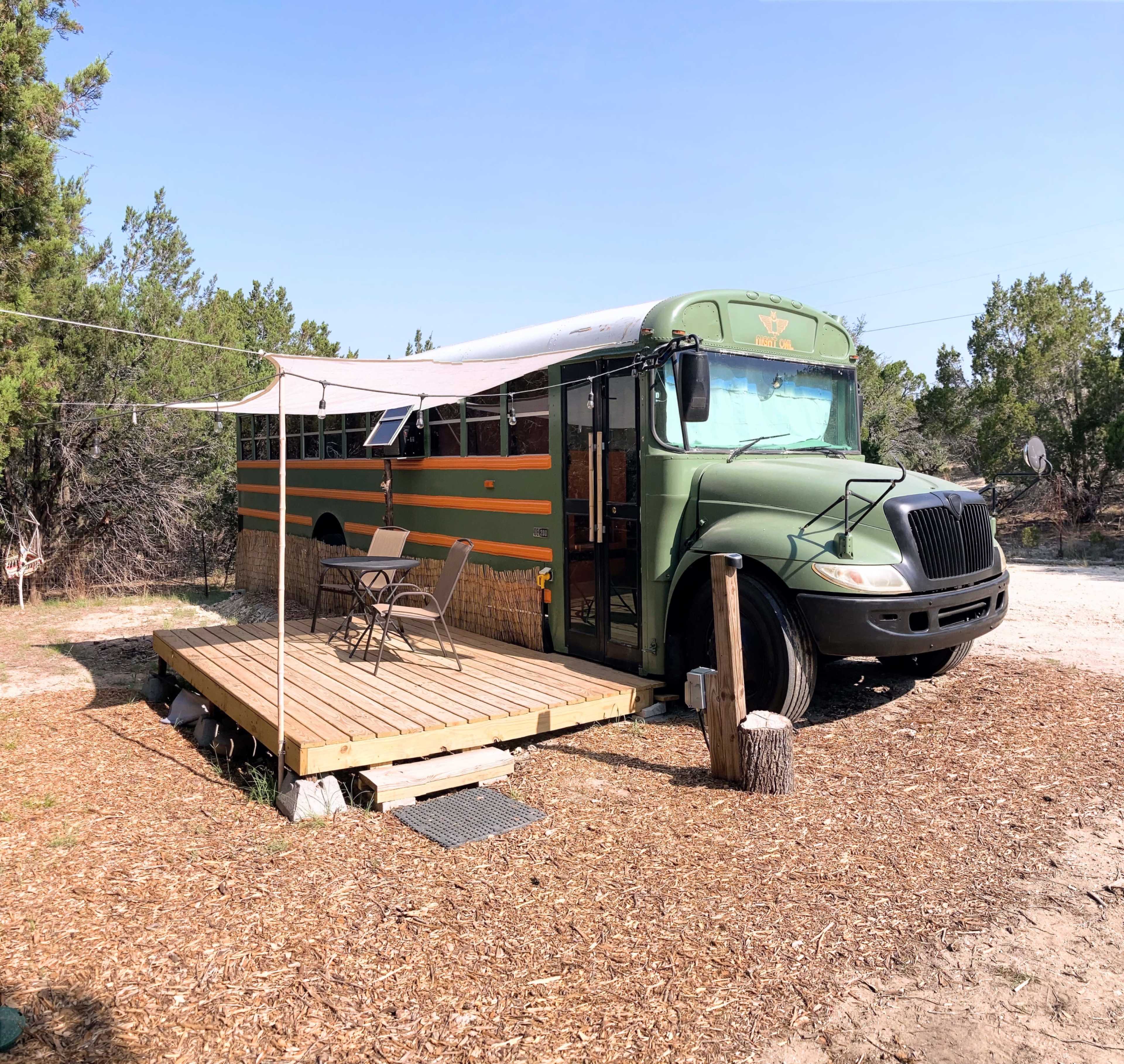 A converted bus is parked next to a wooden deck surrounded by trees in a rural setting.