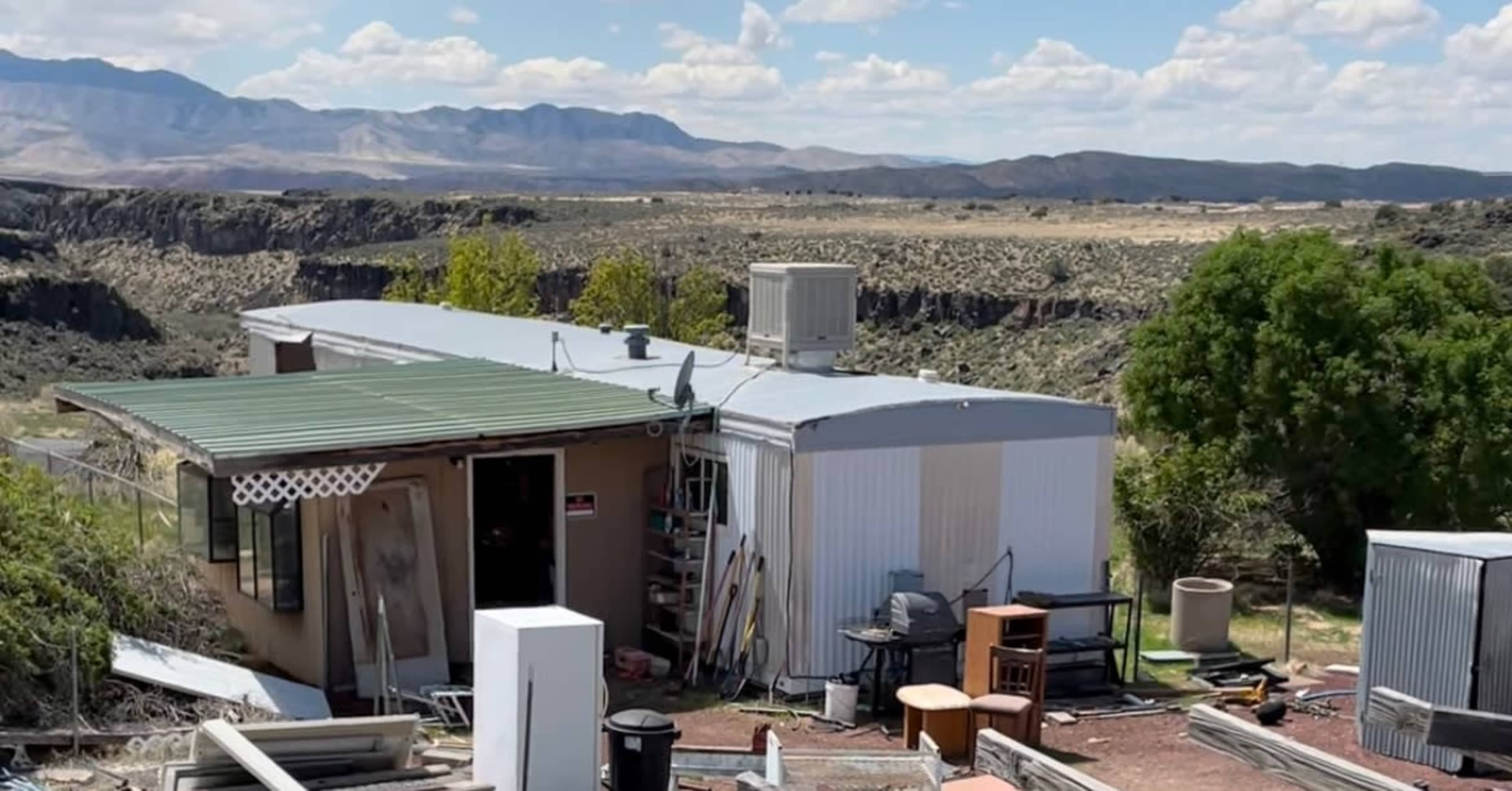 A mobile home sits in a rural landscape surrounded by scattered debris and mountains in the background.