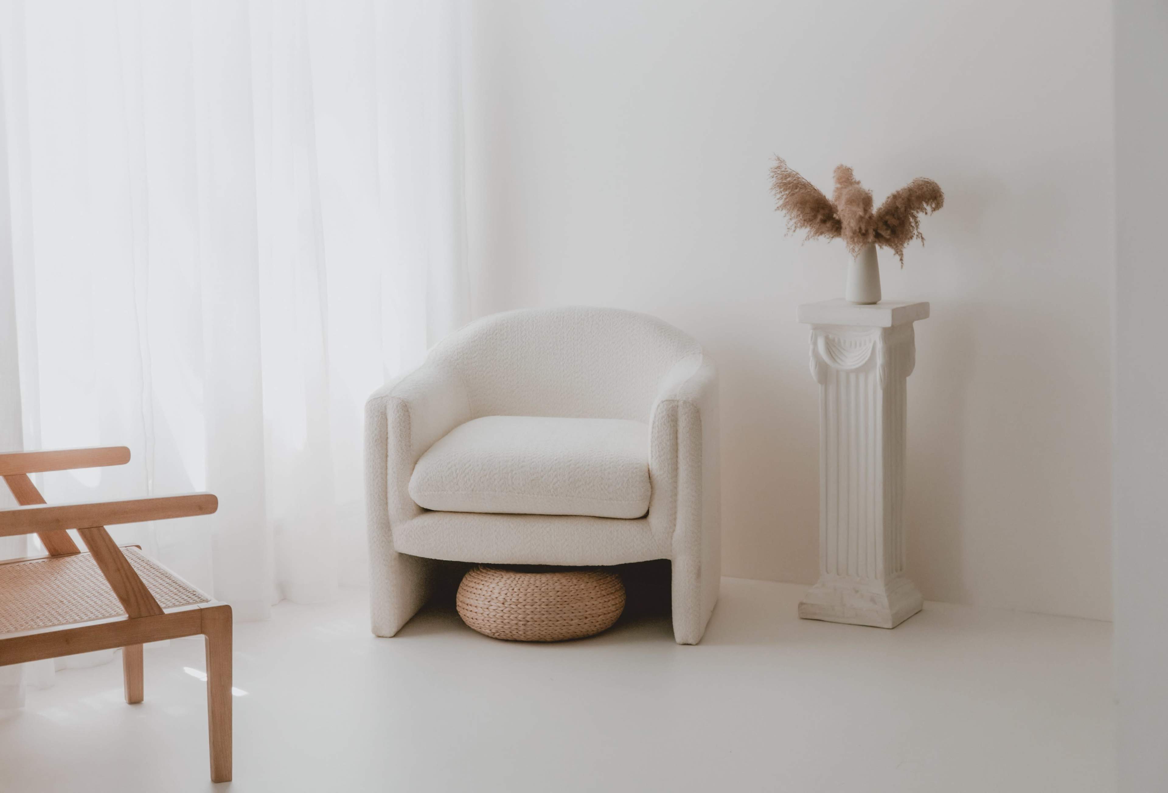 A cozy white armchair is placed next to a decorative column with a vase of pampas grass, set against a bright, minimalist interior.
