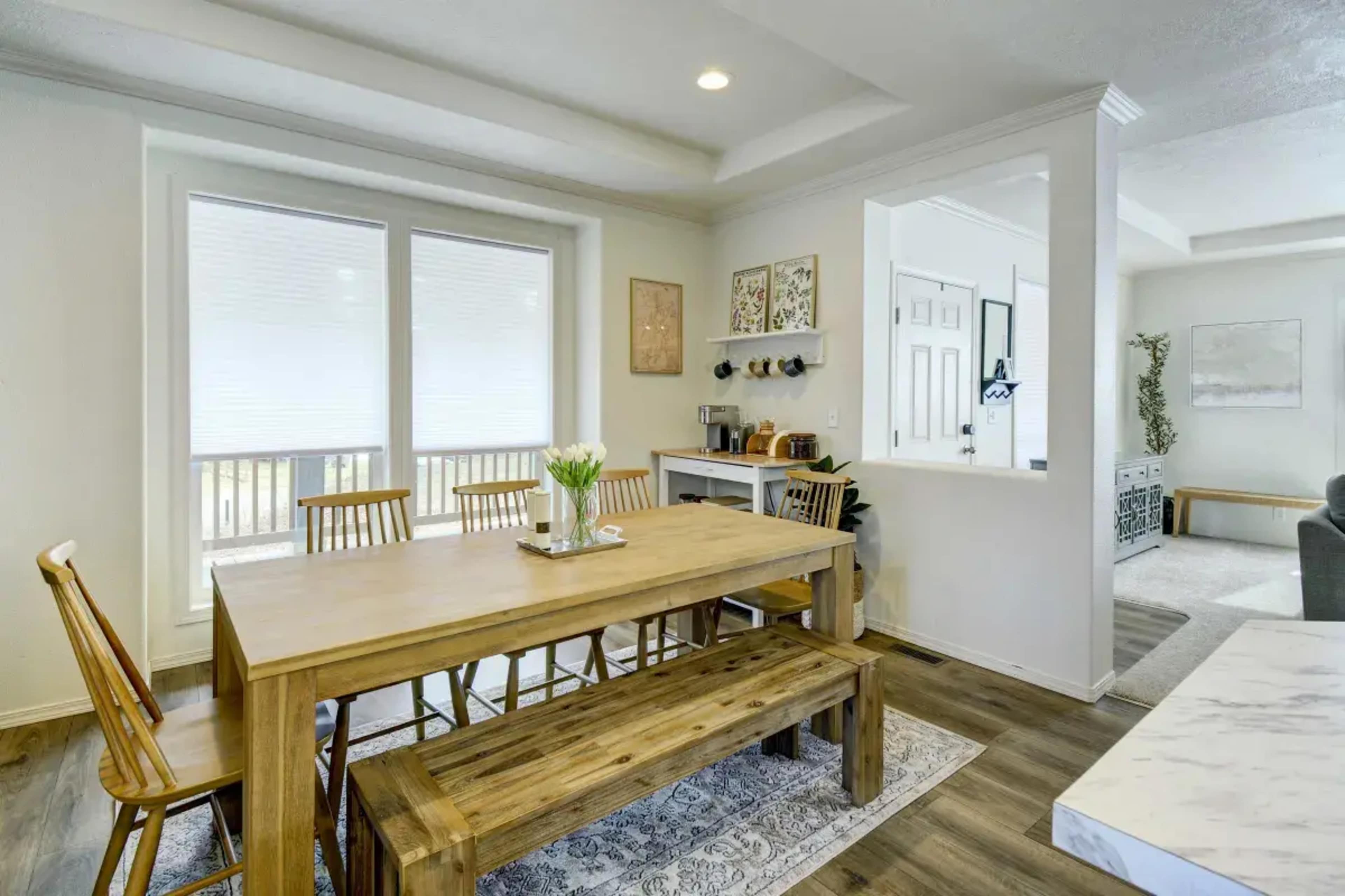 A light-filled dining area featuring a wooden table with chairs and a bench, adjacent to a kitchen counter and a living area in the background.