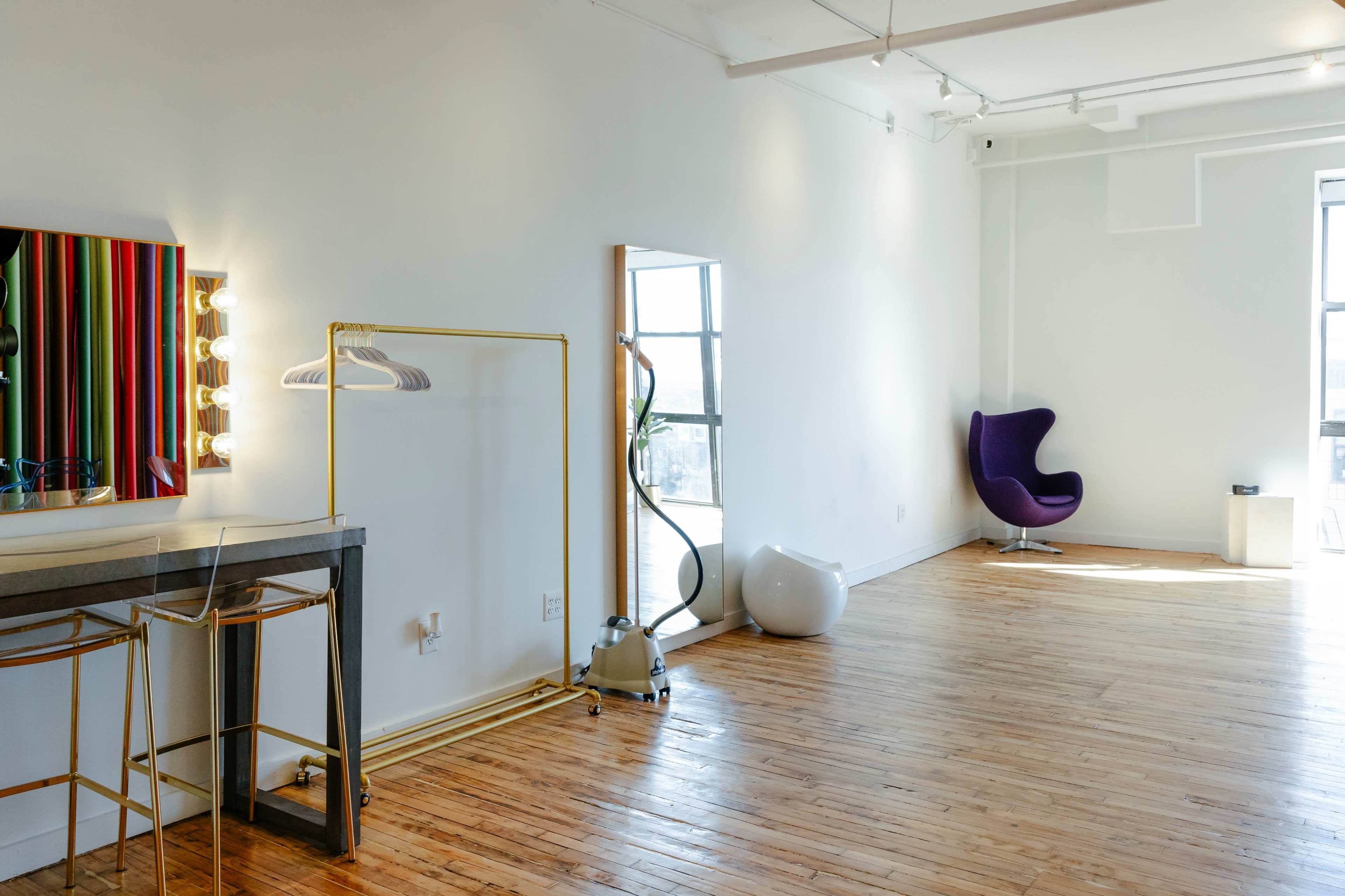 A bright interior space featuring a mirror, a vanity with light bulbs, a purple chair, and wooden flooring.