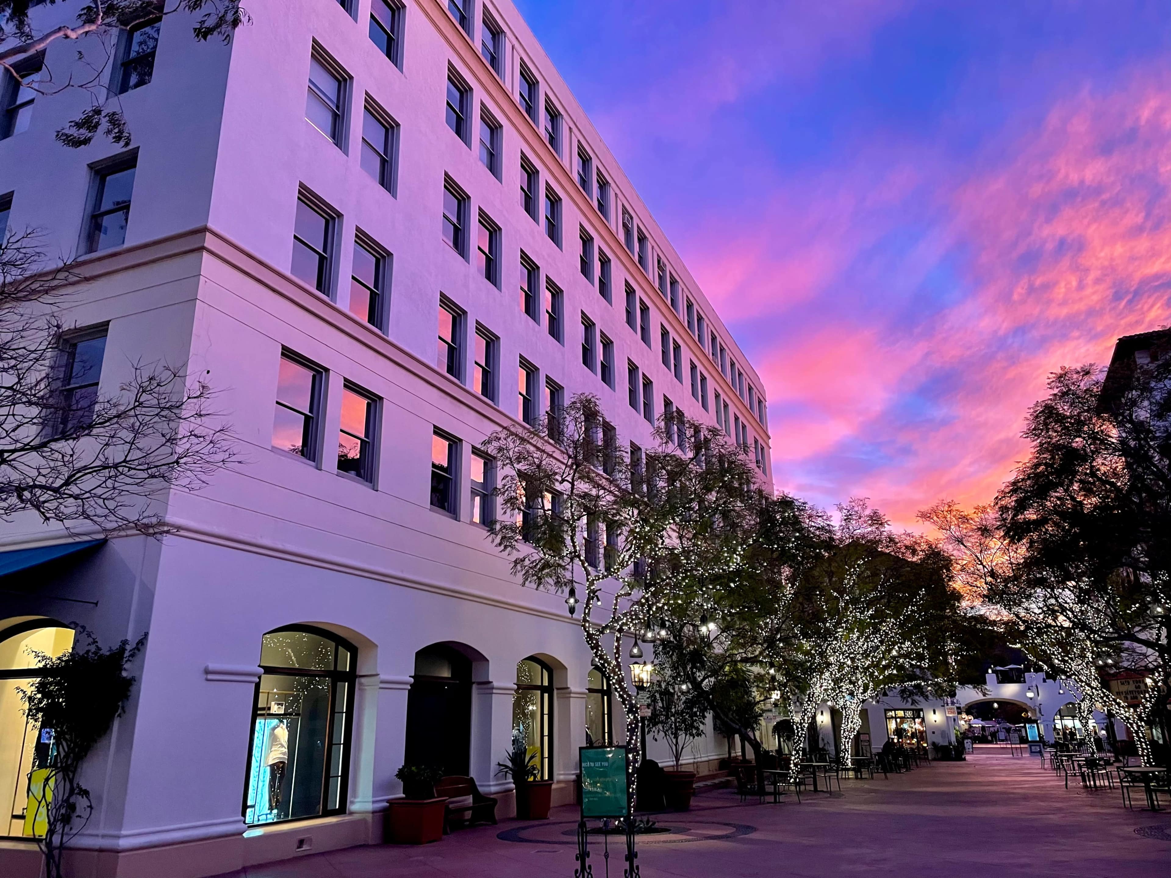 The image shows a wide street lined with a five-story building, illuminated by colorful sunset skies and festive lights on trees.