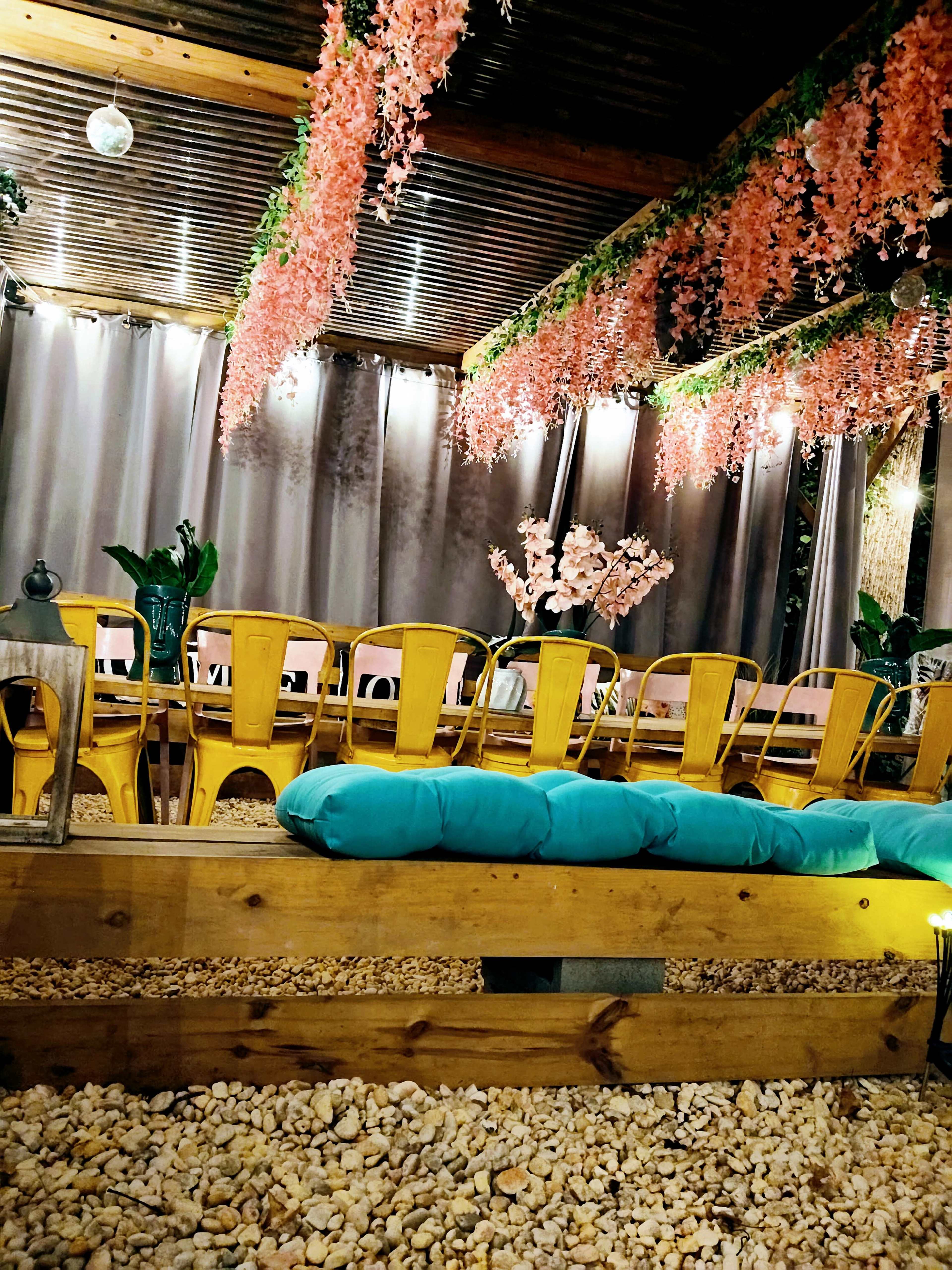 The scene features a cozy indoor space decorated with pink floral arrangements hanging from the ceiling, surrounded by yellow chairs and a wooden bench with blue cushions on a pebbled floor.